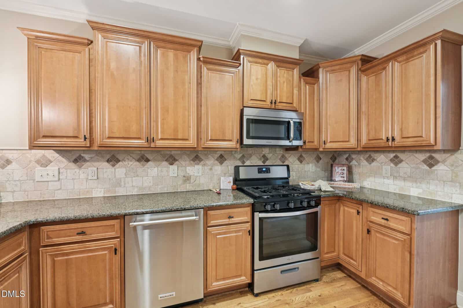 Modern kitchen with light wood cabinets, stainless steel appliances including a gas stove, microwave, and dishwasher, granite countertops, and a beige tile backsplash with a diamond pattern.