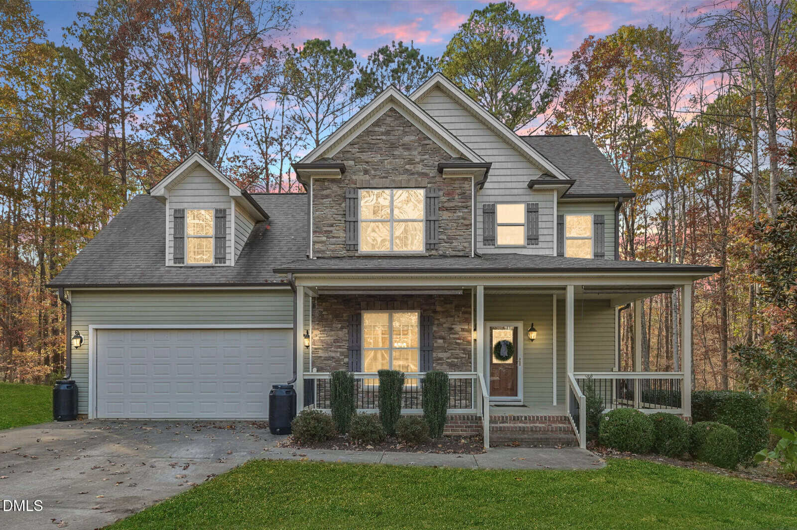 A two-story suburban house with a stone and siding exterior, a covered front porch, an attached two-car garage, and autumn trees in the background at sunset.