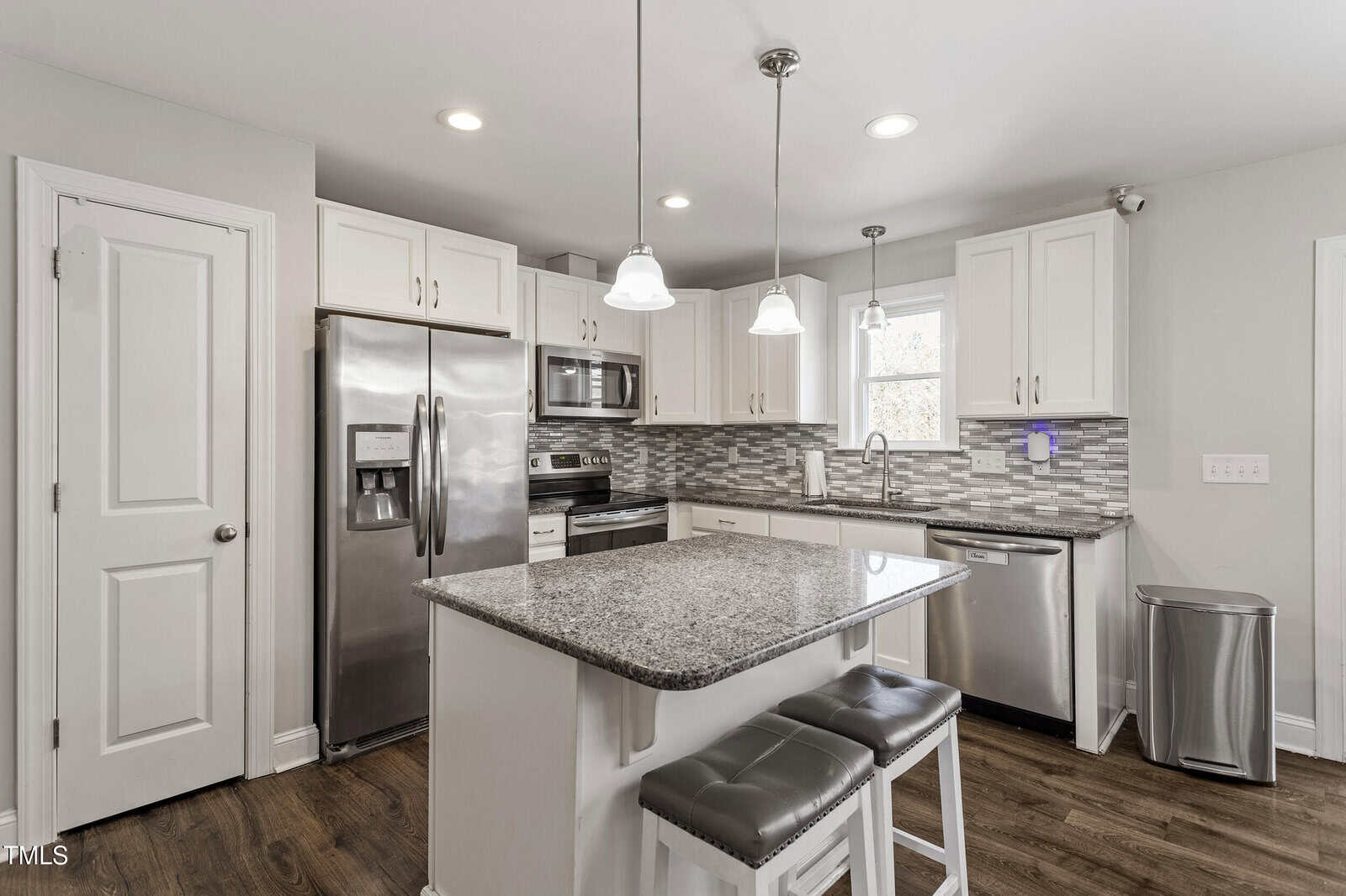 Modern kitchen with white cabinets, stainless steel appliances, granite island, two gray cushioned stools, pendant lights, gray tile backsplash, and wood flooring. Trash can and window are visible.