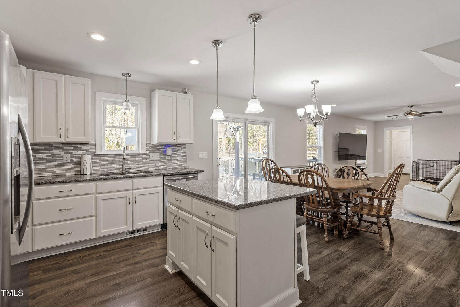 Modern kitchen with white cabinets, granite countertops, island with stools, dining area with wooden table and chairs, pendant and chandelier lights, and wood flooring. Living room area visible in the background.