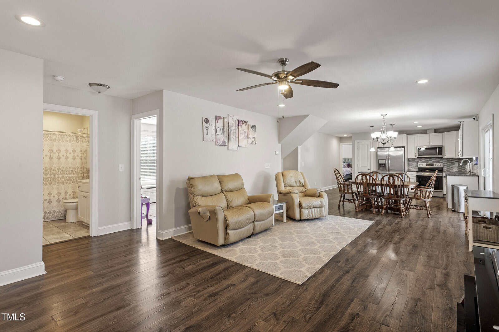 Open living area with two beige recliners on a rug, dining table with chairs, modern kitchen in the background, hardwood floors, ceiling fan, and a glimpse of a bathroom on the left.