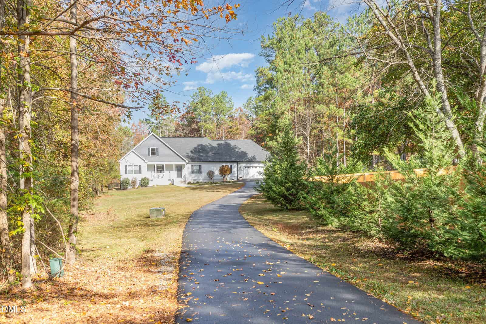 A curved paved driveway leads through a wooded area to a large white house with black shutters and a front lawn, surrounded by trees and greenery on a sunny day.