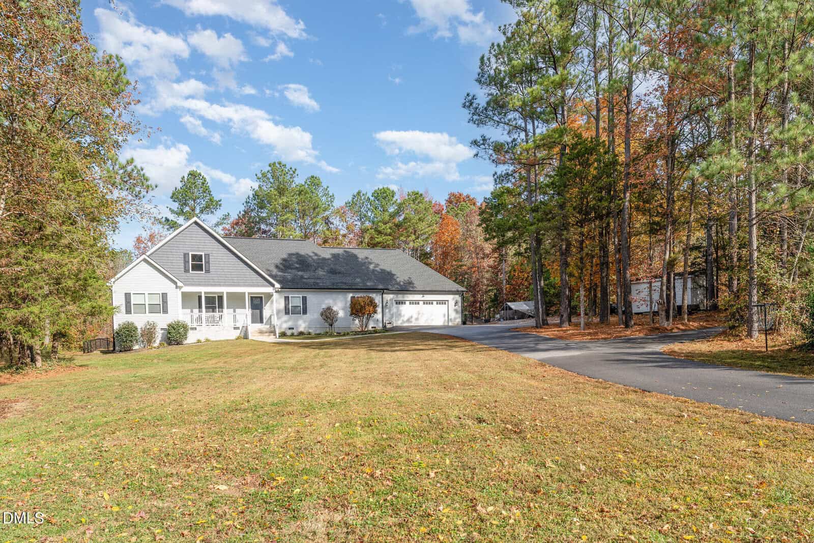 A single-story white house with a large front lawn, gray roof, and attached three-car garage, surrounded by tall trees with autumn foliage under a partly cloudy sky. A paved driveway curves alongside the house.
