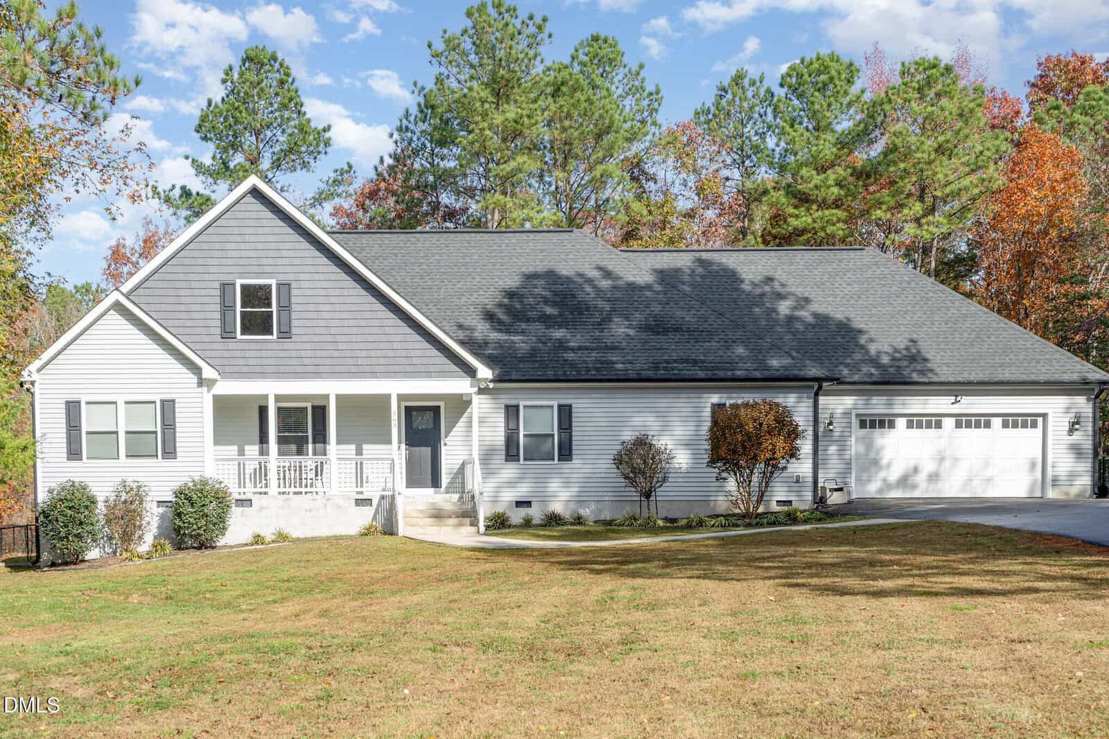 A single-story house with gray siding and a dark gray roof, featuring a front porch, white trim, attached two-car garage, and a lawn with small shrubs and trees. Blue sky and tall trees are in the background.