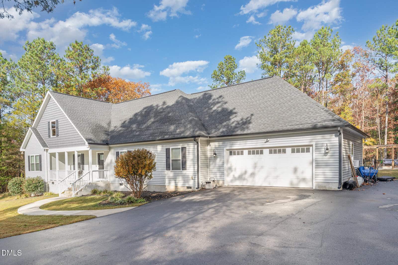 A light gray house with white trim, a front porch, and a two-car garage, surrounded by trees with autumn foliage under a bright blue sky with scattered clouds.