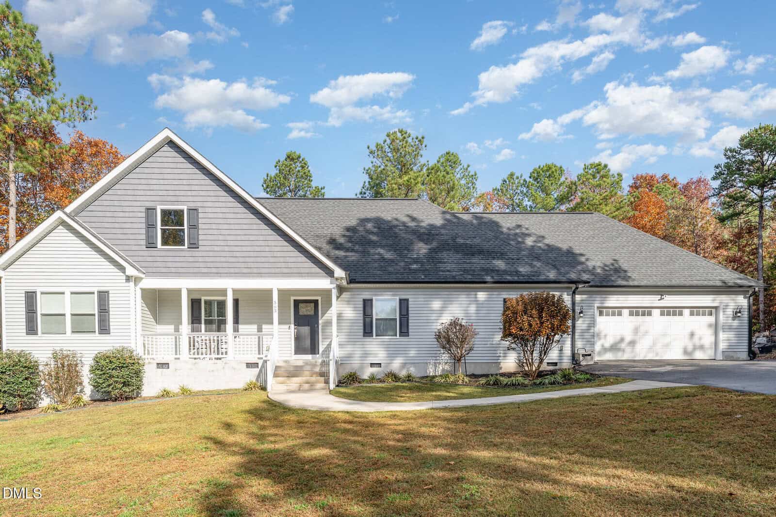 A modern gray and white house with a covered front porch, attached two-car garage, and a well-kept lawn, surrounded by trees with autumn foliage under a partly cloudy sky.