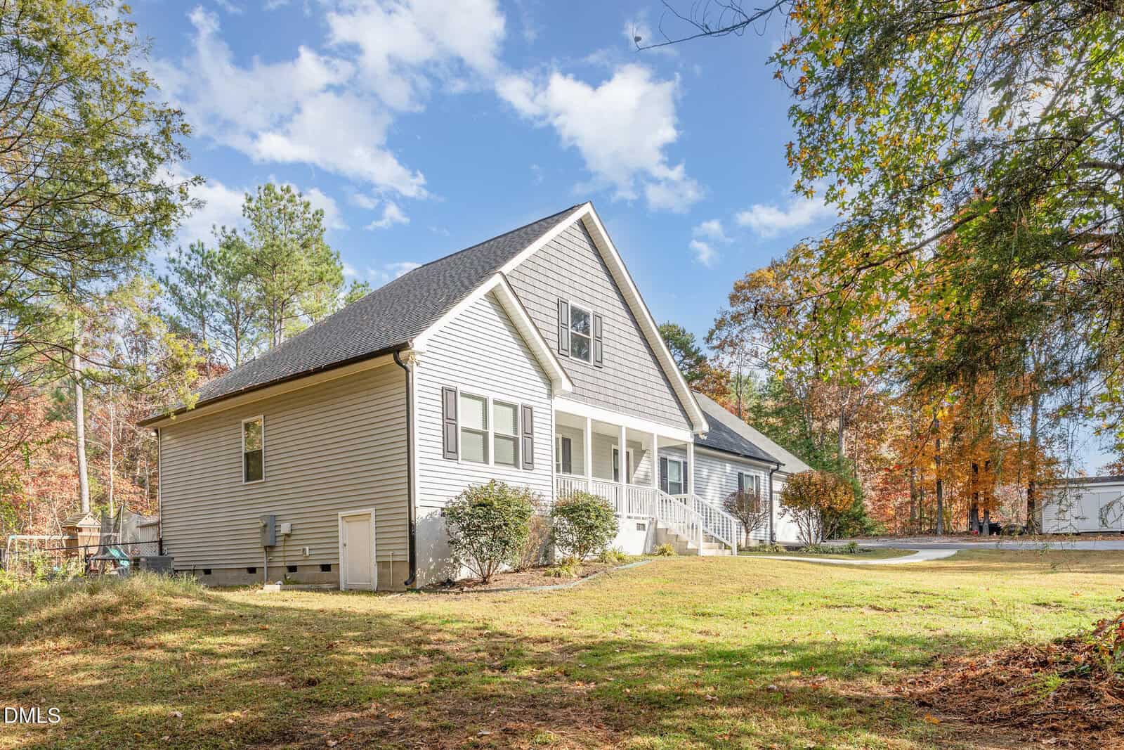 A white single-story house with a dark roof and front porch sits on a grassy yard surrounded by trees with green and autumn-colored leaves under a partly cloudy sky.