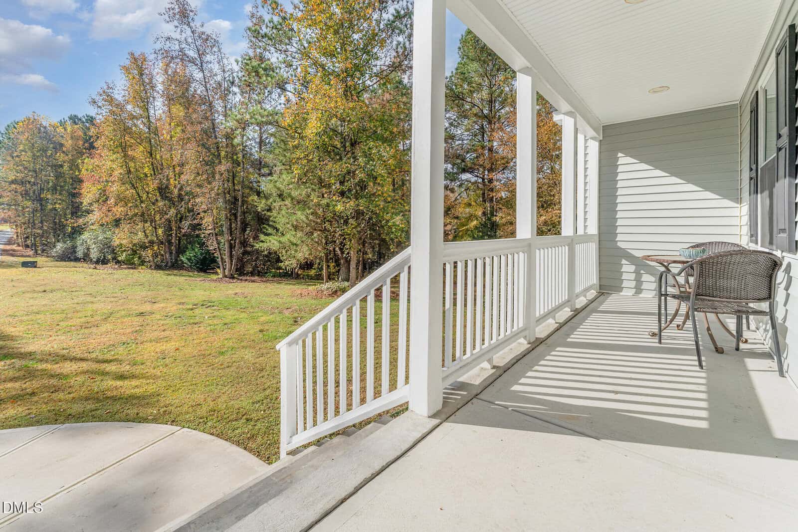 A sunny front porch with white railings, a small round table, and two chairs overlooks a grassy yard bordered by tall trees with autumn foliage.