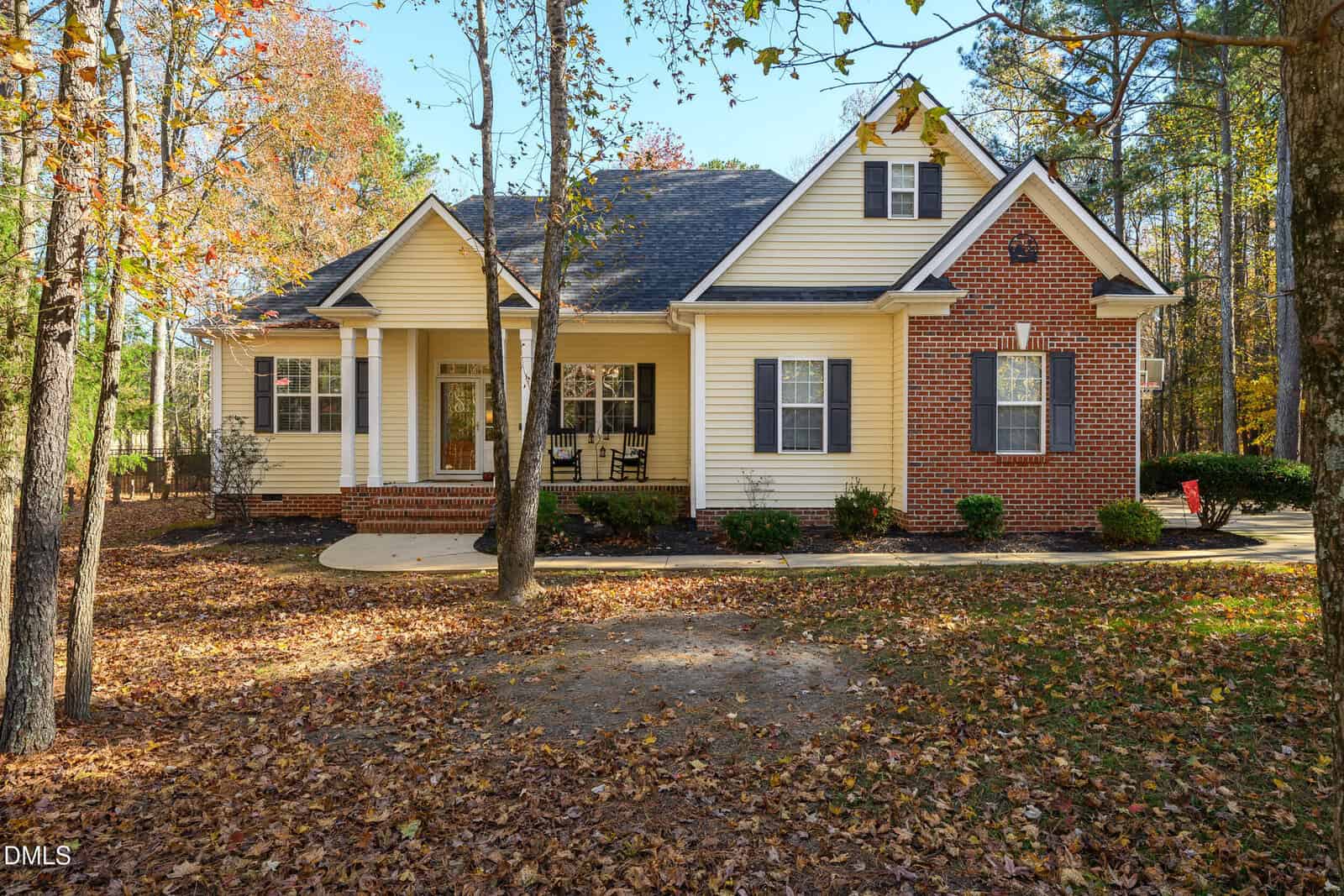 Nestled in a wooded area, 15 Cole Creek Way in Franklinton is a charming single-story house with beige siding, red brick accents, and black shutters. A covered front porch with chairs welcomes you, while mature trees and fallen leaves blanket the front yard. The home also features a classic gable roof, adding to its inviting appeal.