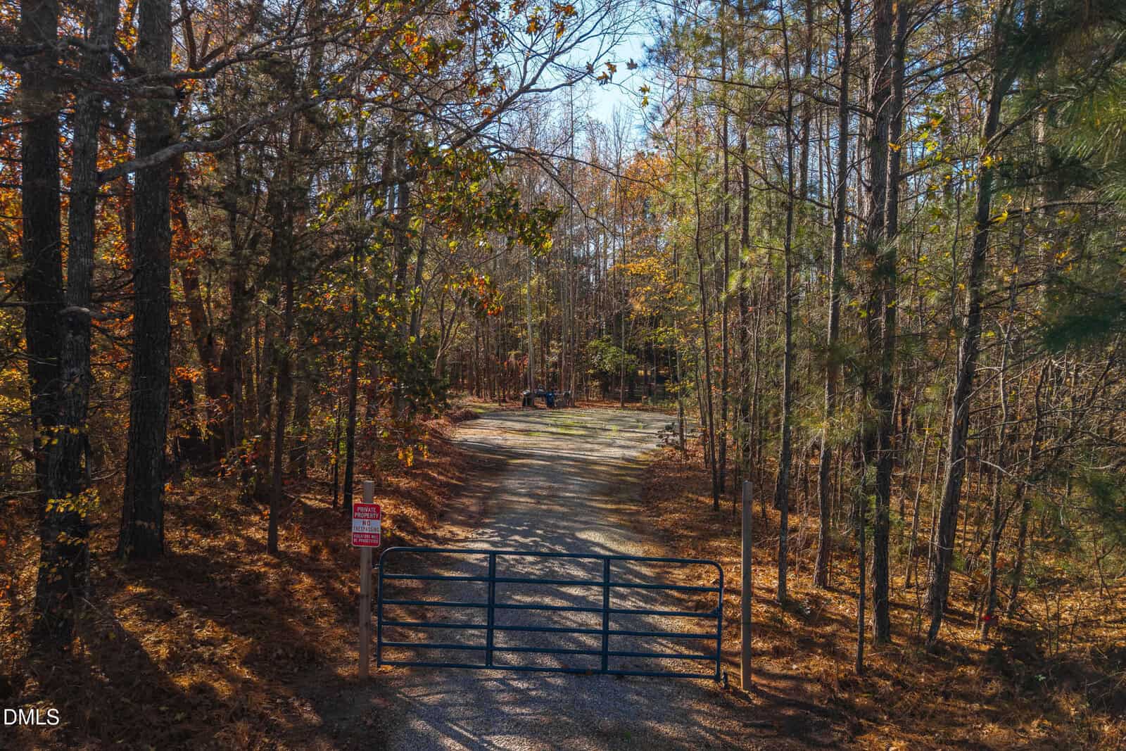A metal gate blocks a gravel road leading into a wooded area with tall trees and autumn foliage. A “No Trespassing” sign is posted on the gate. A car is parked in the clearing in the background.