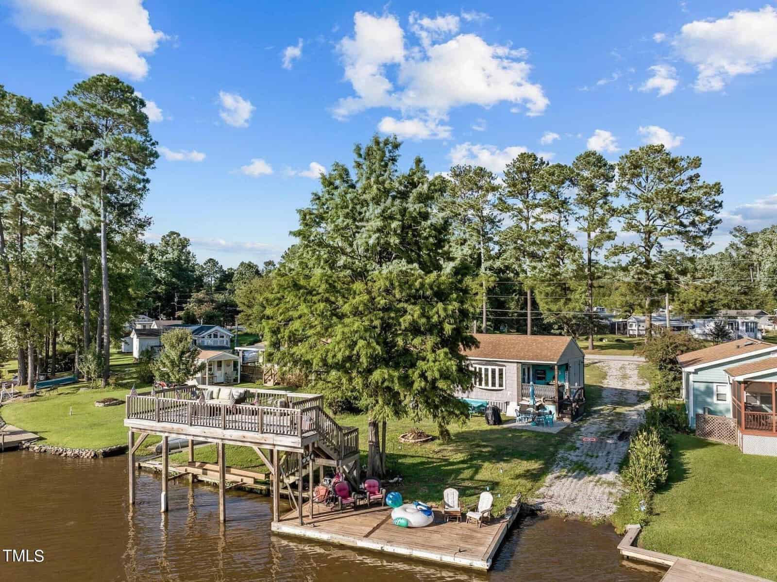 A lakeside property at 1583 Sagamore Drive, Louisburg, featuring a wooden dock and upper deck, several chairs, a fire pit, and a cozy house surrounded by trees. Nearby homes dot the shore beneath a blue sky with scattered clouds.