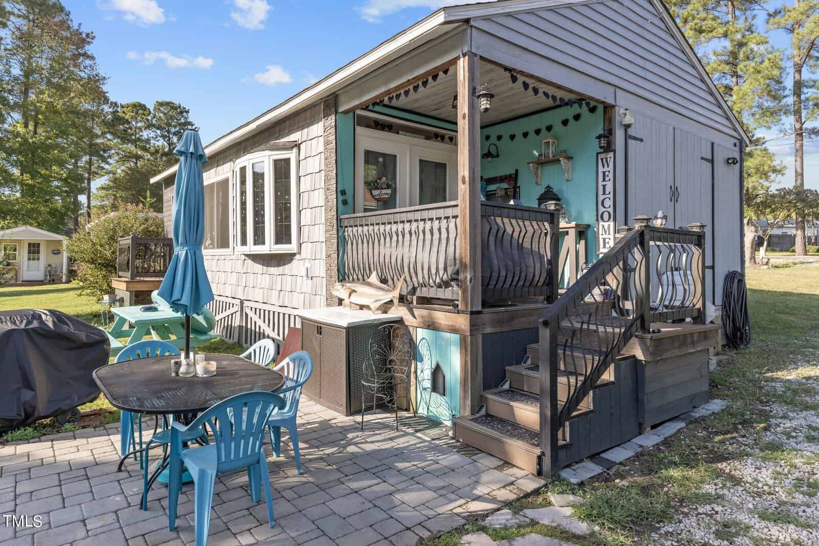 A small gray house with a covered porch displays a "Welcome" sign by the stairs. The yard features a patio area with a blue table, matching chairs, and a large umbrella next to a grill on a stone paver surface.