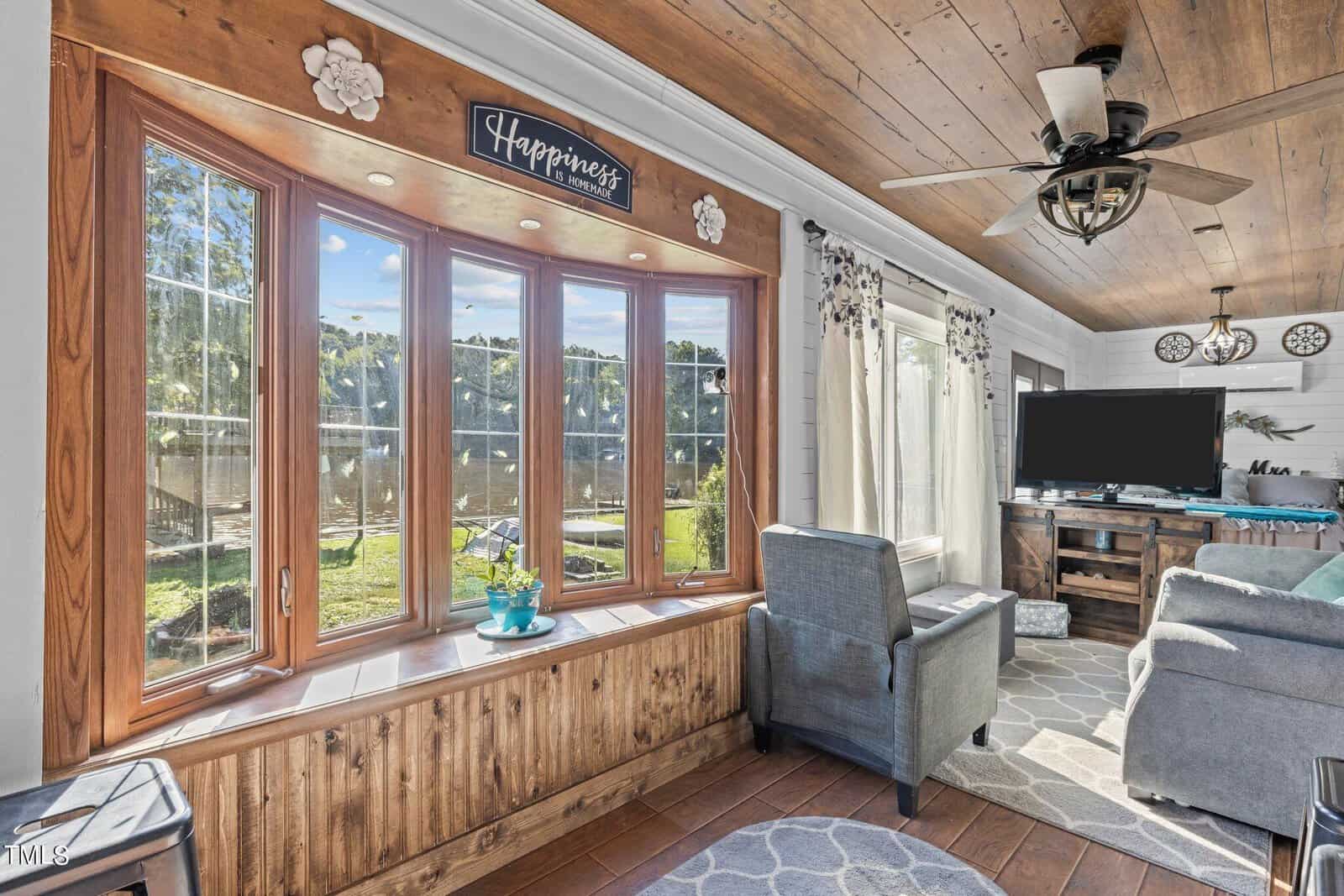 Sunroom with large bay window, wood ceiling, gray armchairs, a rug, and a ceiling fan. Sunlight streams in, highlighting the “Happiness” sign above the window and a view of trees and a yard outside.