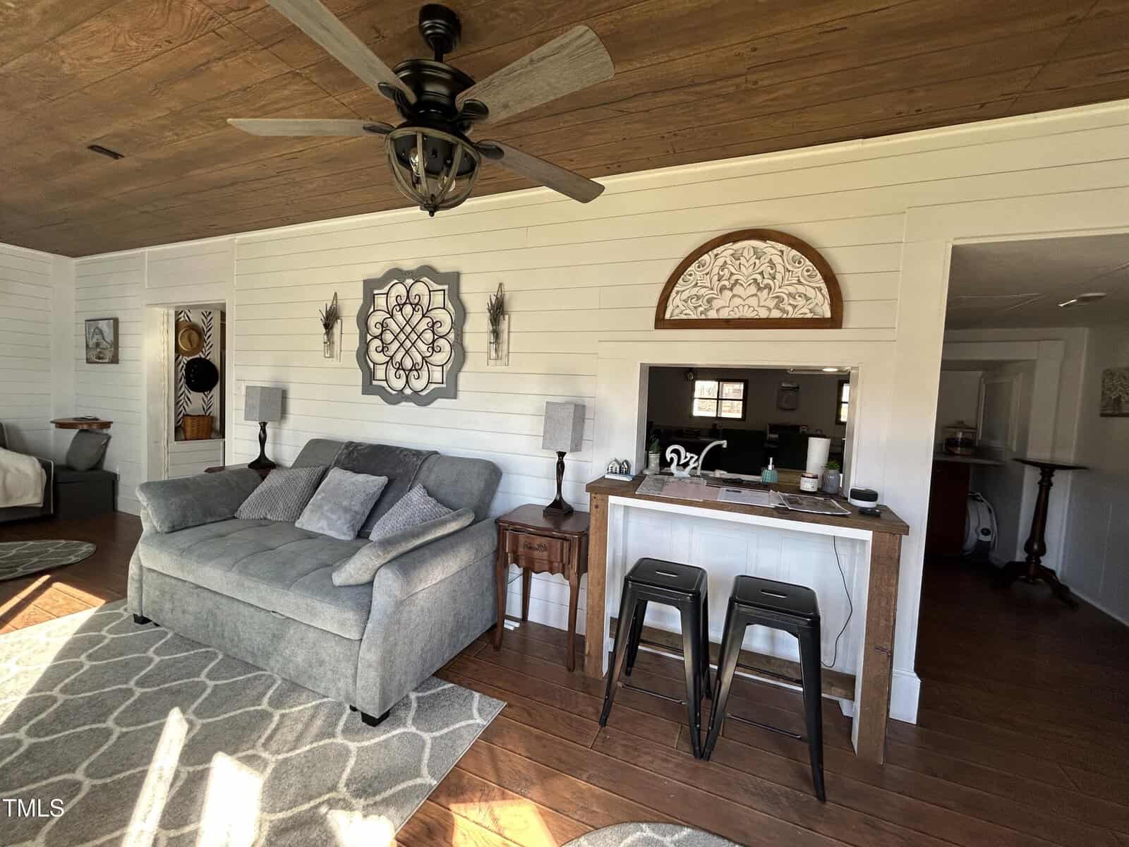 A living room with a gray couch, two side tables with lamps, a wall-mounted decorative piece, ceiling fan, wooden ceiling, bar with two black stools, and a patterned rug on a wooden floor.