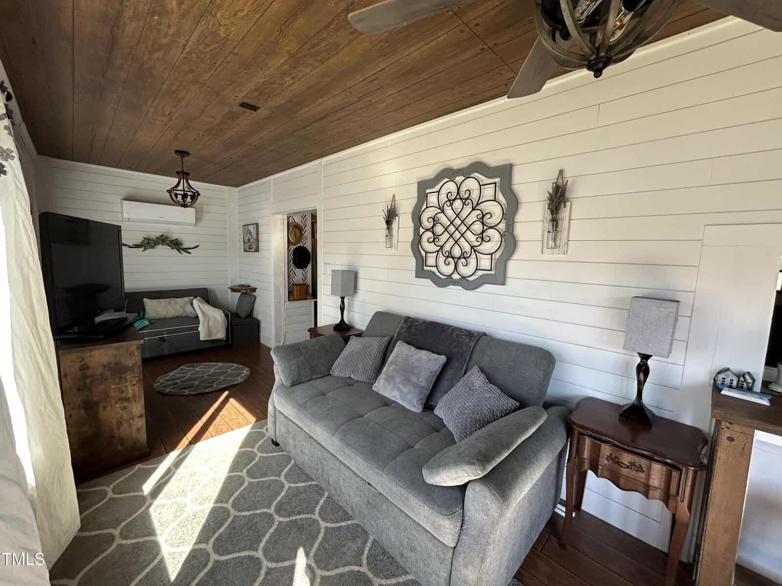A living room with a gray couch, patterned rug, wooden ceiling, white shiplap walls, two side tables with lamps, a TV, and a daybed in the corner. Sunlight enters through a window on the left.