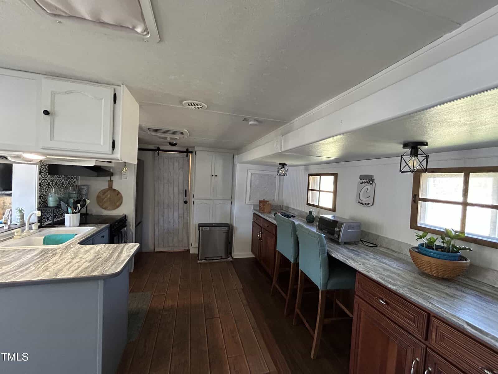 A kitchen with white upper cabinets, dark lower cabinets, a marble-pattern countertop, a sink, and a stove. There is a wooden floor, a long desk with two teal chairs, windows, and various kitchen items.