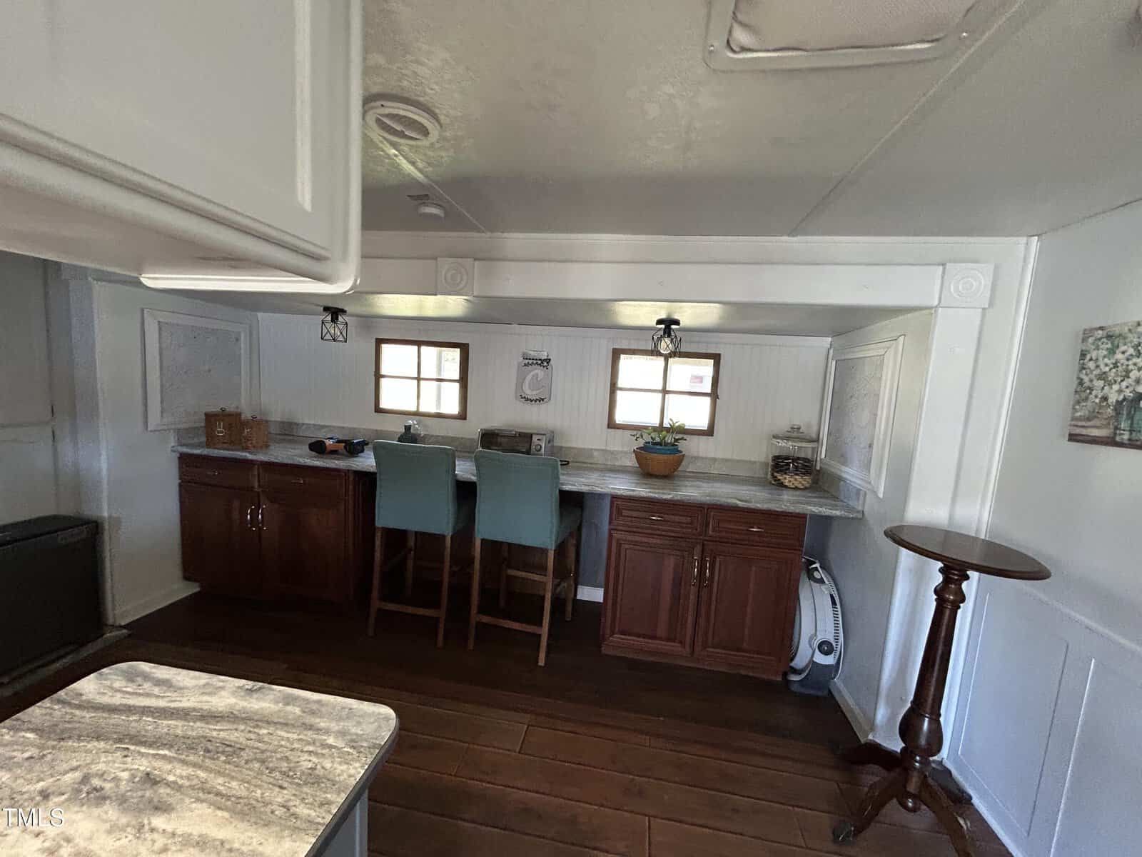 A small kitchen area with dark wooden cabinets, granite countertops, two light blue bar stools, small windows, a potted plant, and a round wooden side table on a dark wood floor.