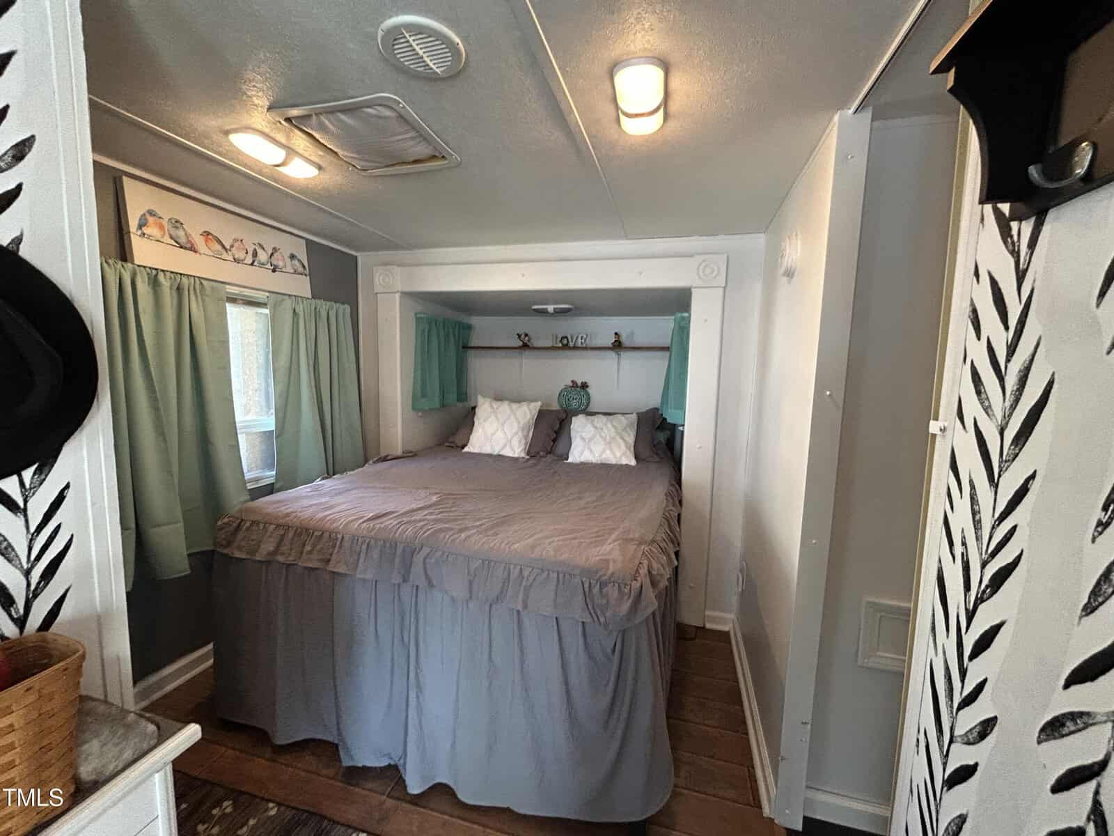 A small bedroom with a queen-sized bed covered in gray bedding, flanked by two green curtains and windows. The walls are gray and white with black leaf patterns, and there is wooden flooring.