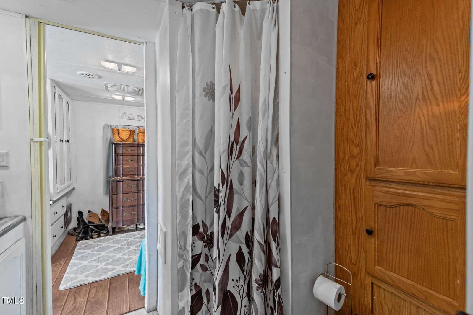 A small bathroom features a floral-patterned shower curtain, a wooden cabinet with a toilet paper roll holder, and a view into an adjoining room with a rug, dresser, and shoes near the wall.
