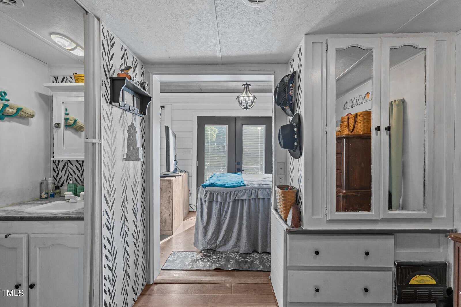 A view from a hallway into a bedroom with double doors at the far end. The hallway features cabinets, drawers, and a mirror on the right, and a bathroom with a sink and decorative wallpaper on the left. The floors are wood.