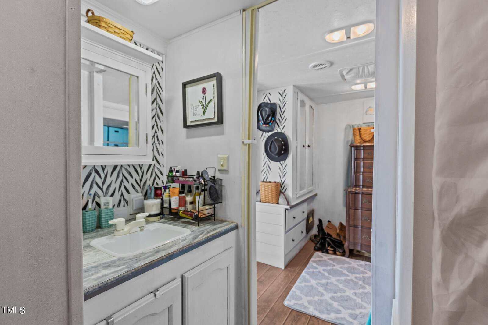 A small bathroom with a sink, mirror, and toiletries on the counter. The wall has a black-and-white herringbone pattern. Through the doorway, a dresser, shelves, hats, shoes, and a rug are visible in the adjoining room.