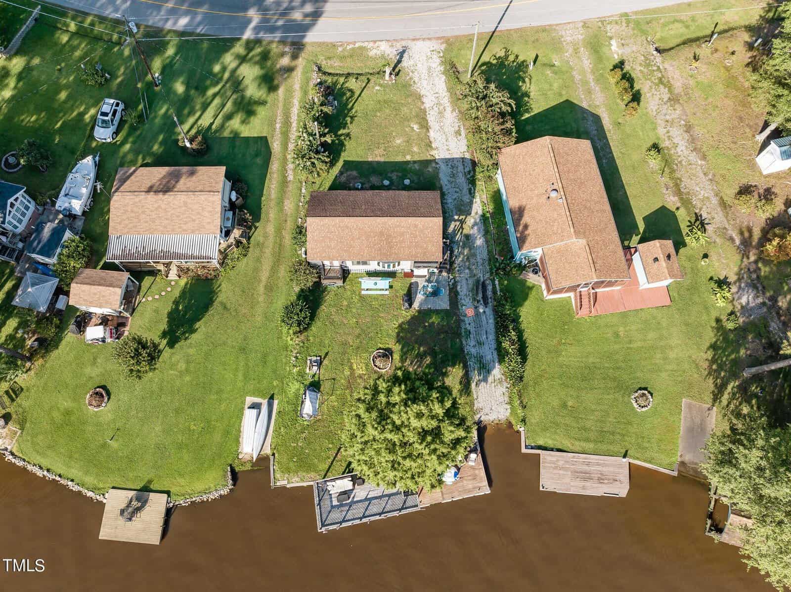 Aerial view of three houses with brown roofs by a road and a river, each with green lawns, docks, trees, and boats in their yards.