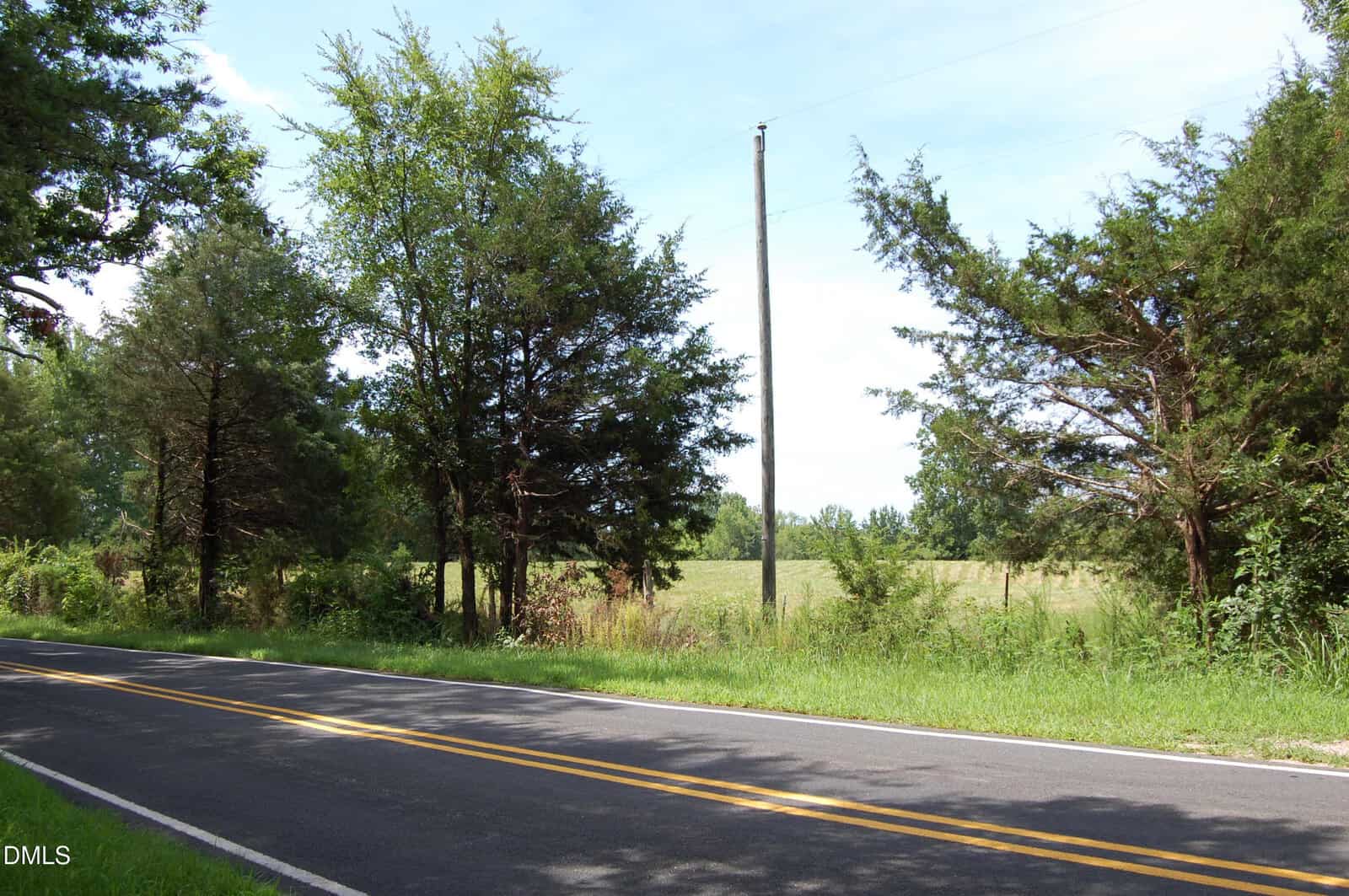 A paved two-lane road with double yellow lines runs beside grassy land and trees on a clear day. Power lines and a utility pole are visible in the background.