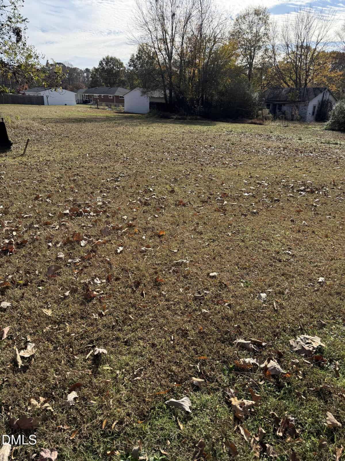 A grassy, mostly flat vacant lot with scattered leaves, bordered by trees and a few small houses or sheds in the background under a partly cloudy sky.