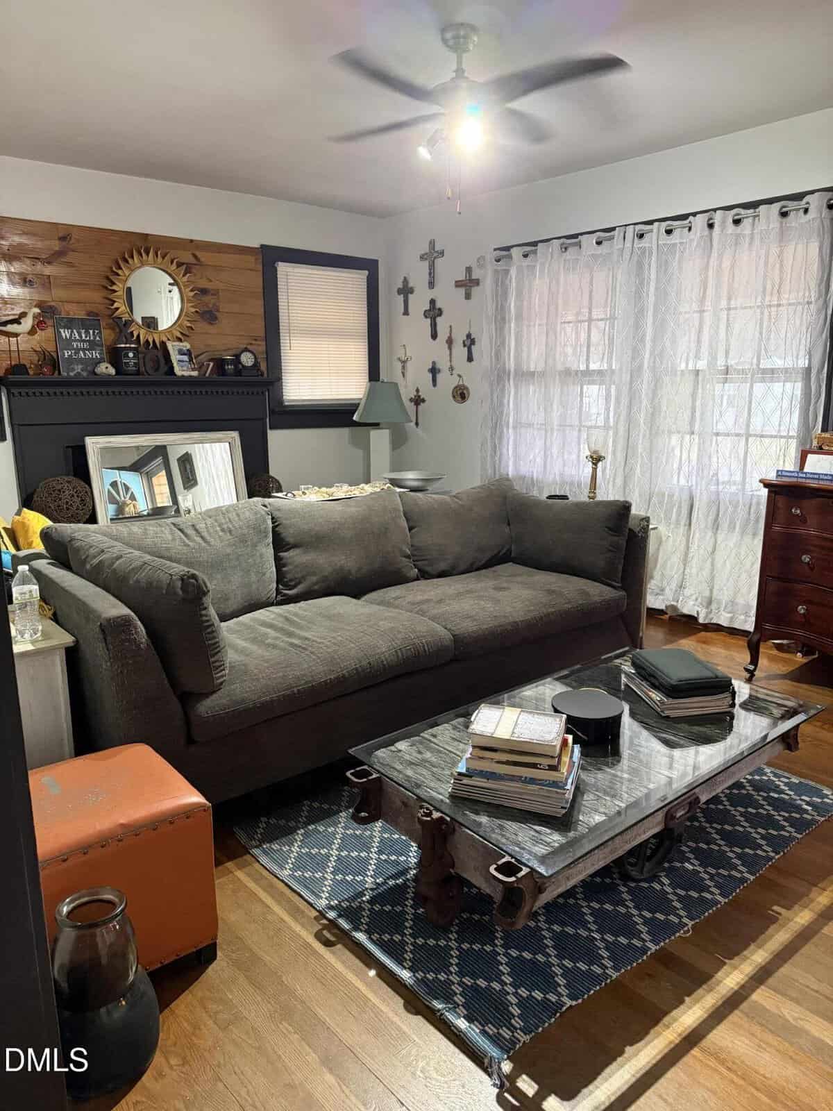 Living room with a gray couch, glass coffee table, books, wall with crosses, sheer white curtains, hardwood floor, patterned rug, and various decor items on tables and shelves. Ceiling fan with light is on.