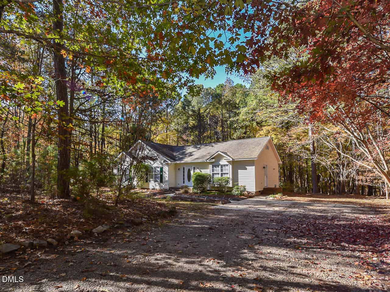A single-story white house with a gray roof sits surrounded by trees with autumn foliage. The gravel driveway leads up to the house, and sunlight filters through the colorful leaves.