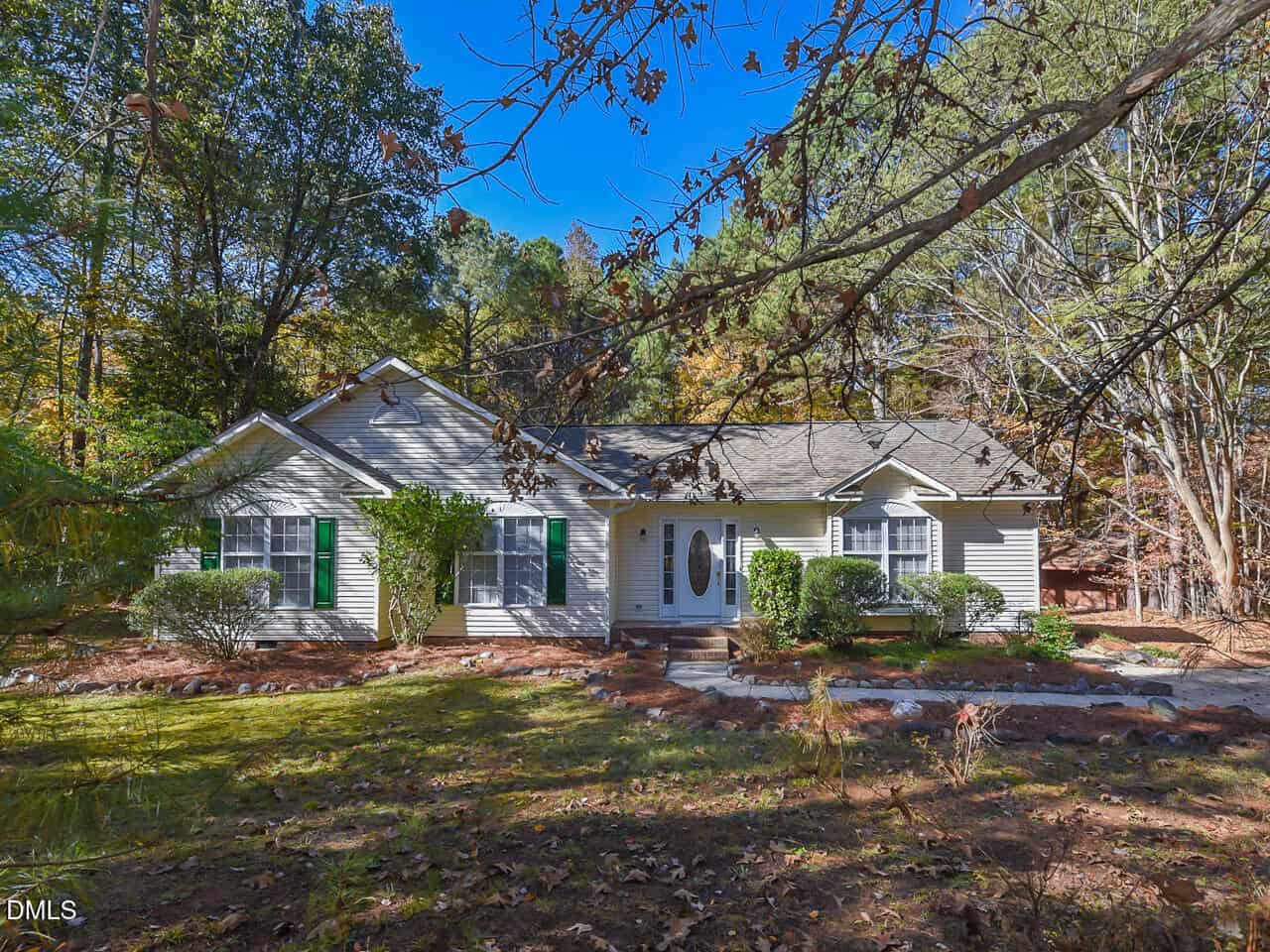 Charming single-story house at 720 Timberlands Drive, Louisburg, with light-colored siding and green shutters, surrounded by trees and shrubs. A clear blue sky brightens the scene, while fallen leaves cover the front yard.