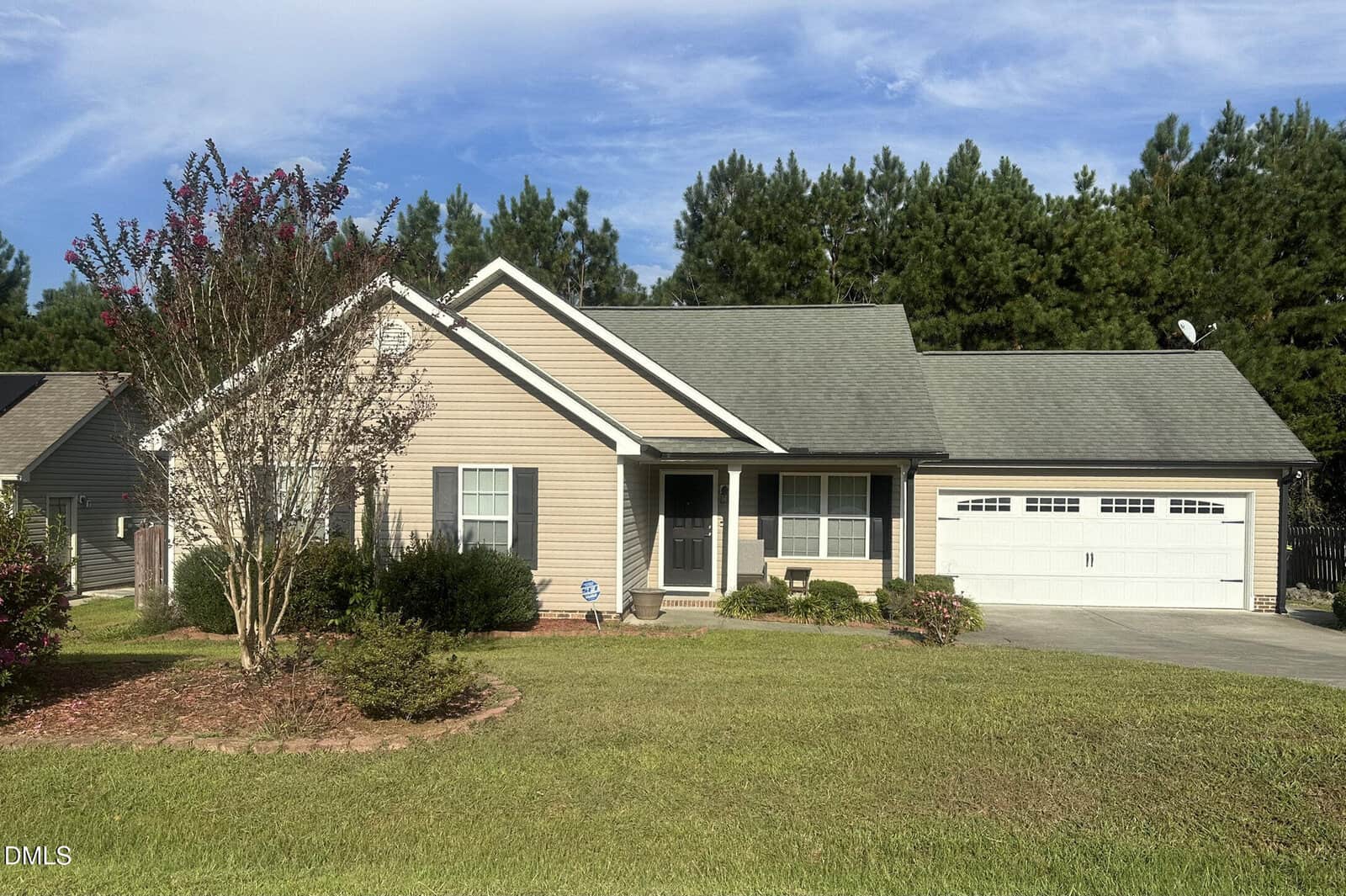 Single-story beige house with white trim, attached two-car garage, grey roof, and a small front porch. Front yard has grass, shrubs, and a tree. Pine trees and a partly cloudy sky are visible in the background.