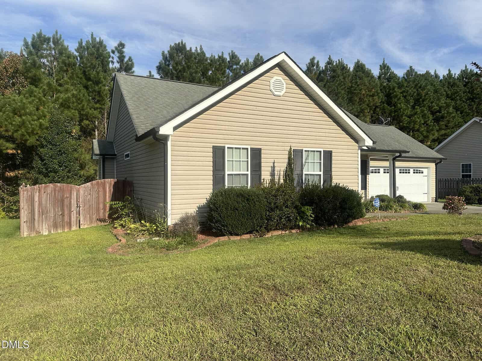 A single-story beige house with white trim, gray shingles, and two front windows. There is a garage on the right and a wooden fence on the left. The yard is grassy with some bushes by the house and trees in the background.