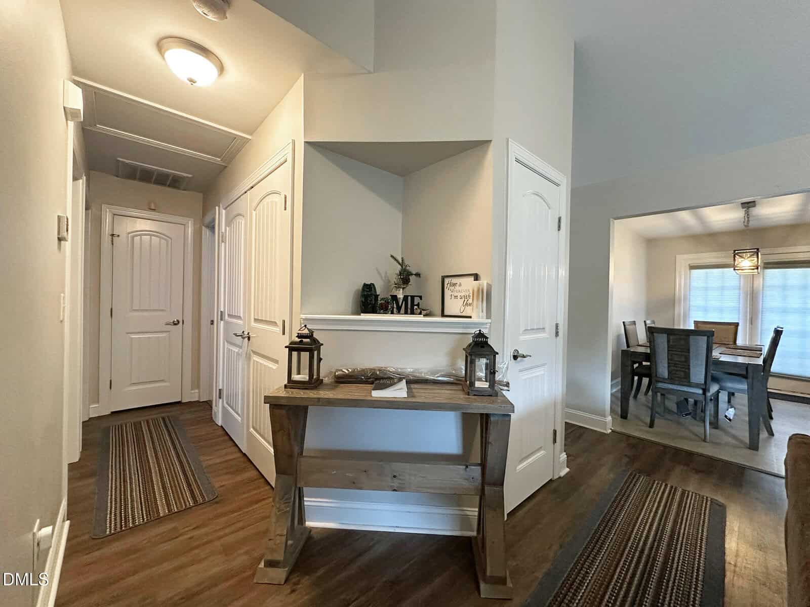 A hallway in a home with white doors, wood flooring, and a wooden console table decorated with lanterns and small items. An adjacent dining room with a table and chairs is visible to the right.