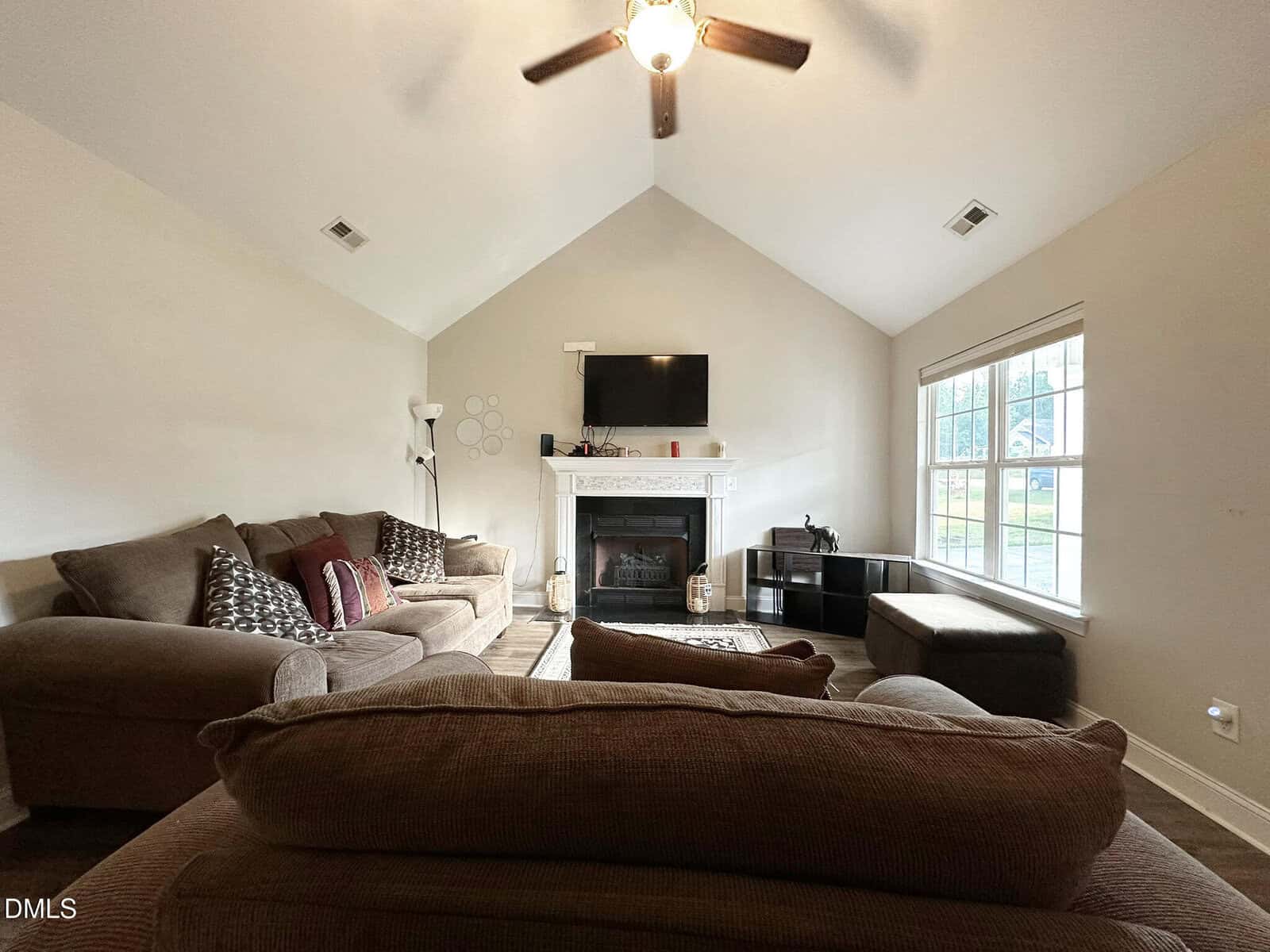 A living room with a brown sectional sofa, a fireplace topped with a TV, a lamp, decorative pillows, a large window letting in daylight, and a ceiling fan on the white vaulted ceiling.