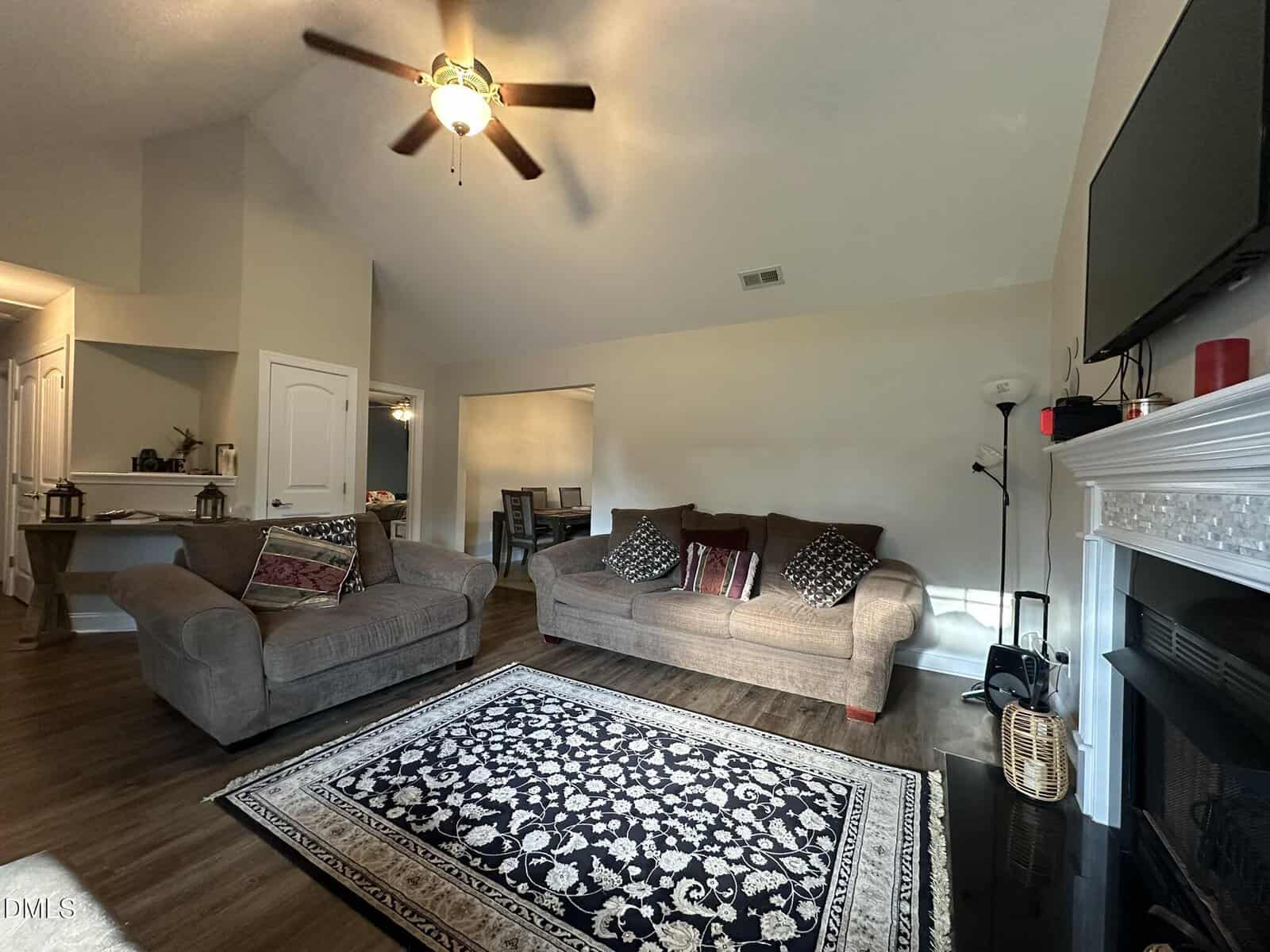 A living room with a ceiling fan, two sofas, patterned rug, and a fireplace. A TV is mounted above the fireplace. There are side tables, lamps, and decorative cushions. The dining area is visible through an open doorway.