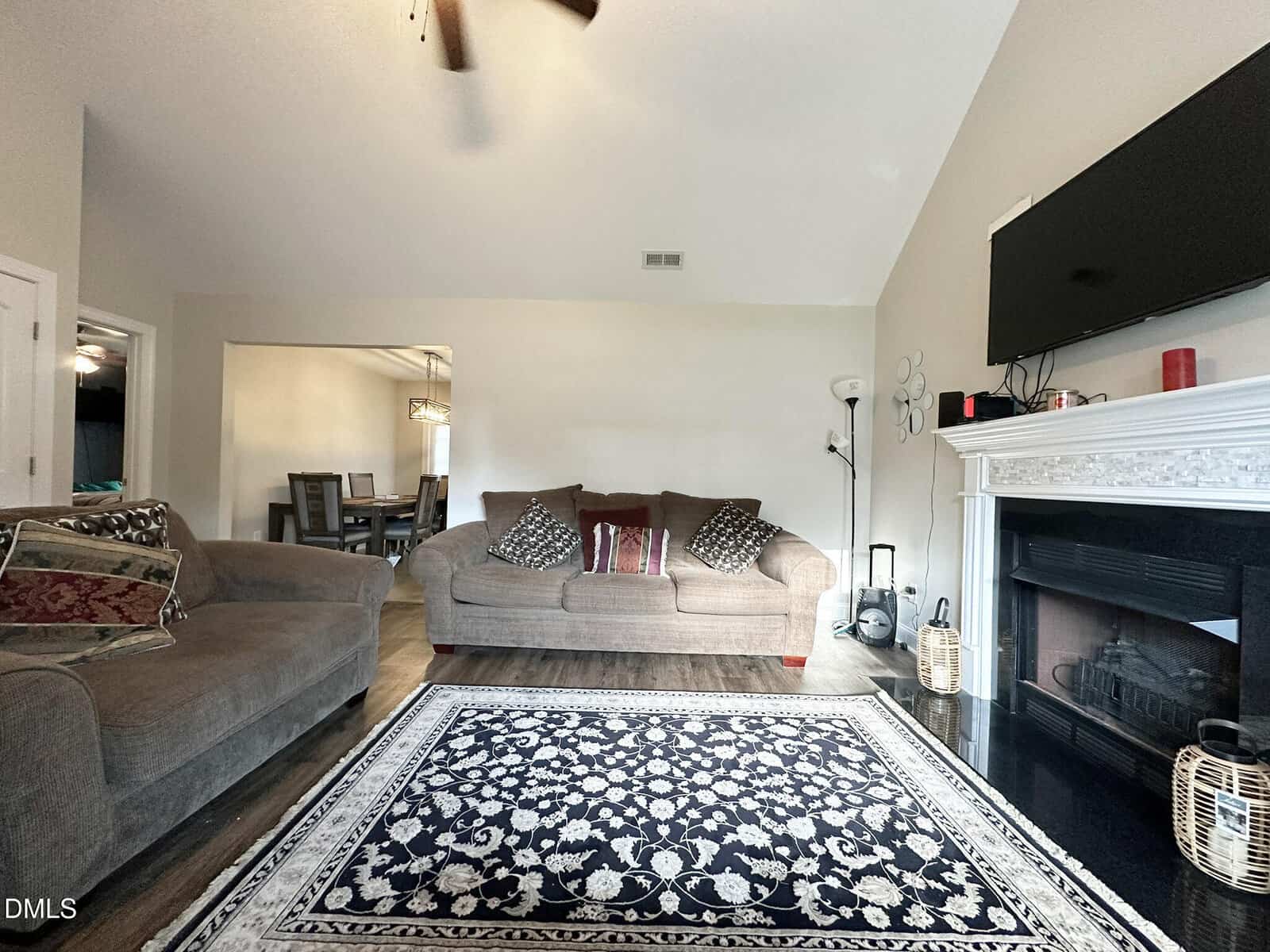 A cozy living room with two sofas, a patterned rug, a wall-mounted TV above a white fireplace, and a dining area visible in the background. The space is warmly lit and neatly arranged.