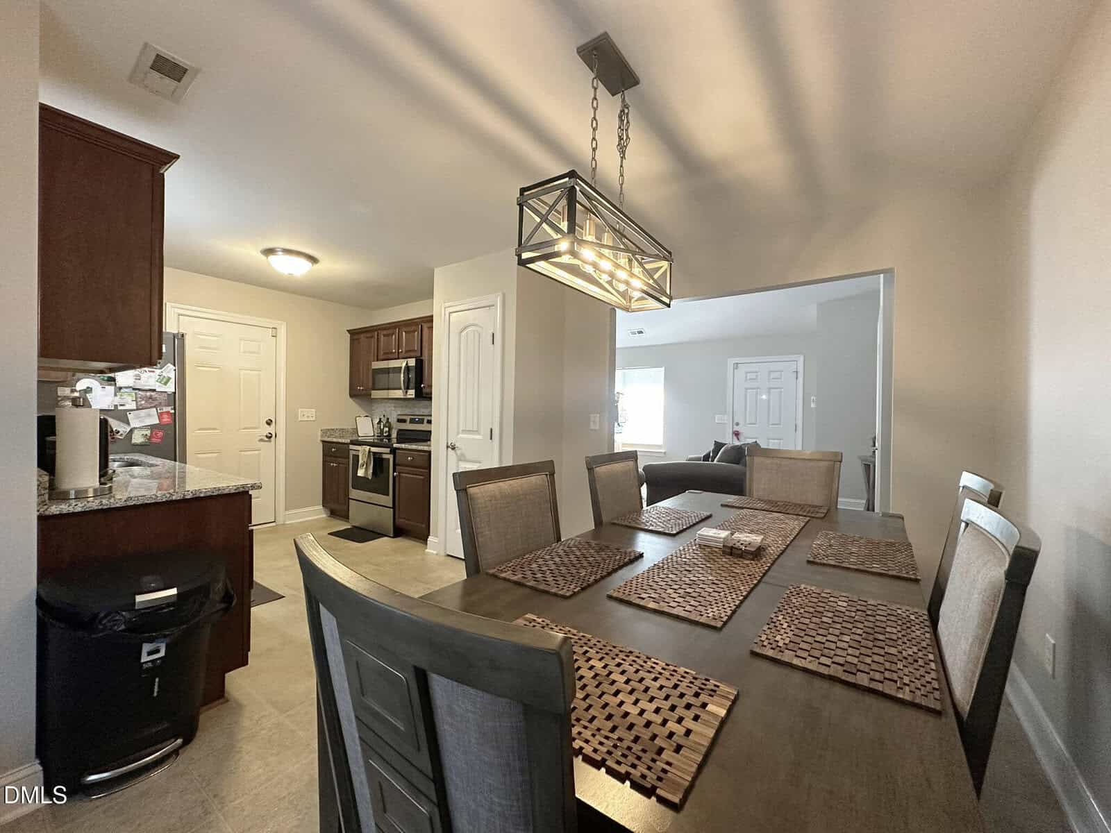 A dining room with a dark wooden table set for eight, modern light fixture above, kitchen in the background with stainless steel appliances, and a living room visible through a wide doorway.