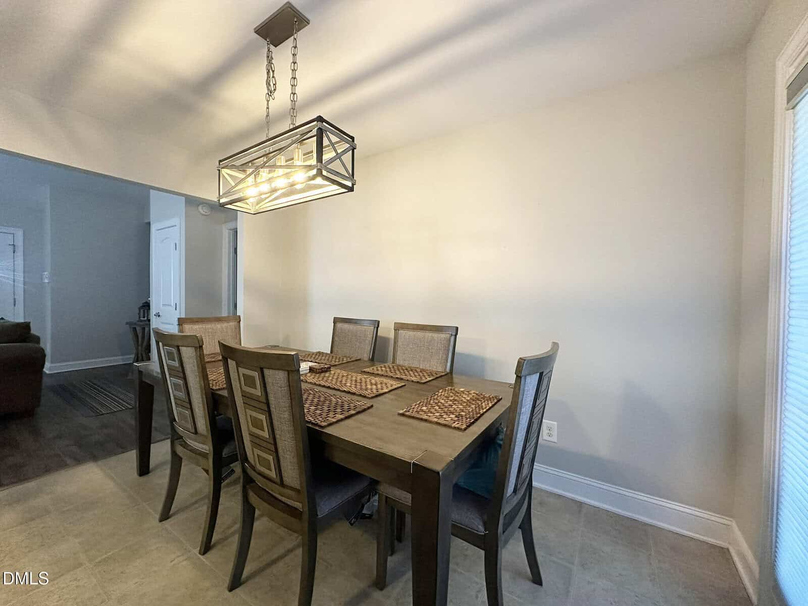 A dining room with a rectangular wooden table, six chairs, woven placemats, and a modern rectangular chandelier. The walls are light-colored, and sunlight enters through a window on the right.
