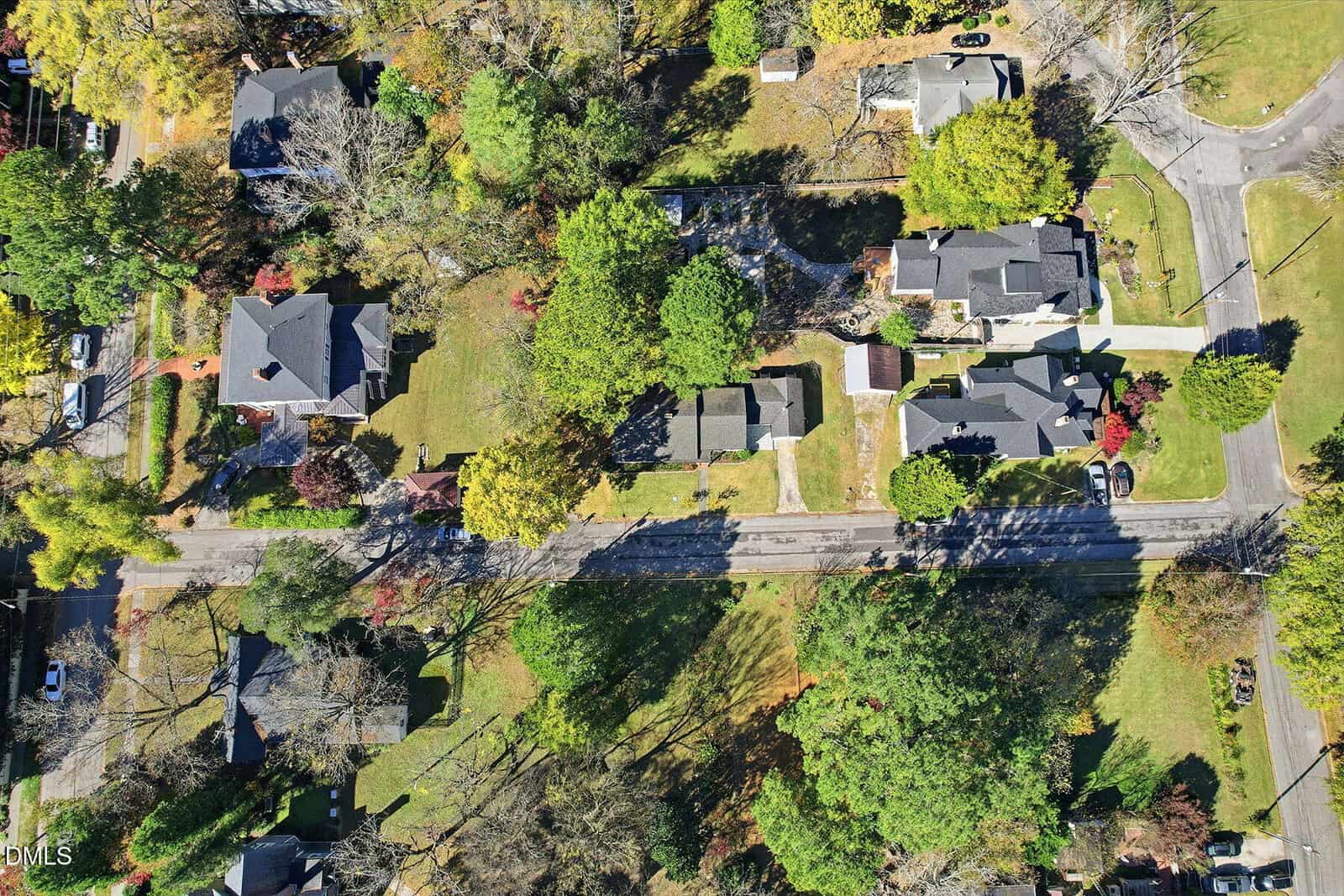 Aerial view of a suburban neighborhood with several houses, green lawns, trees, and a two-lane road running horizontally through the center. Cars are parked in driveways and along the street.