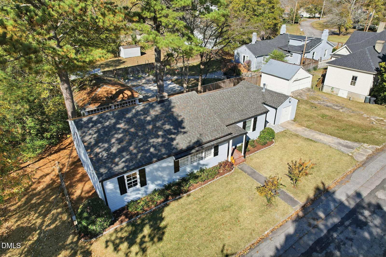 Aerial view of a single-story house with a gray roof and white exterior, surrounded by a grassy yard with a driveway and trees casting shadows. Neighboring houses and a street are visible in the background.