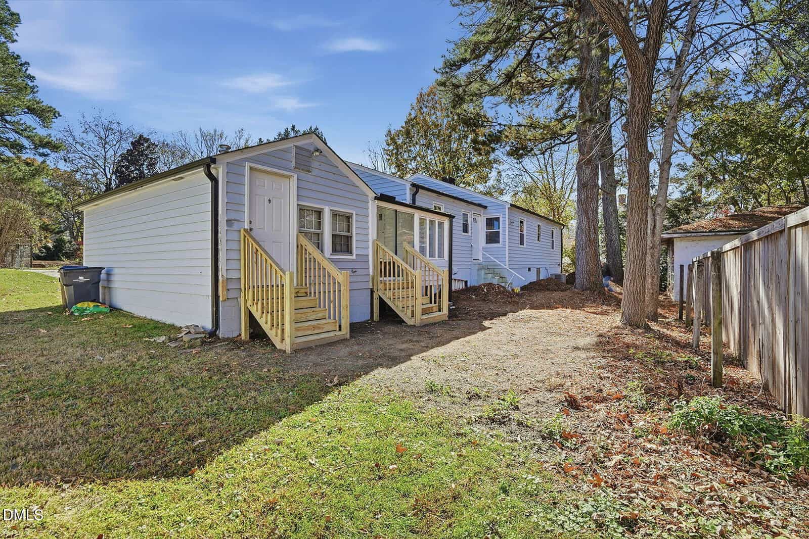 Small white house with two wooden porches and steps, surrounded by grass, trees, and a wooden fence on the right under a blue sky. Trash bin is next to the house.