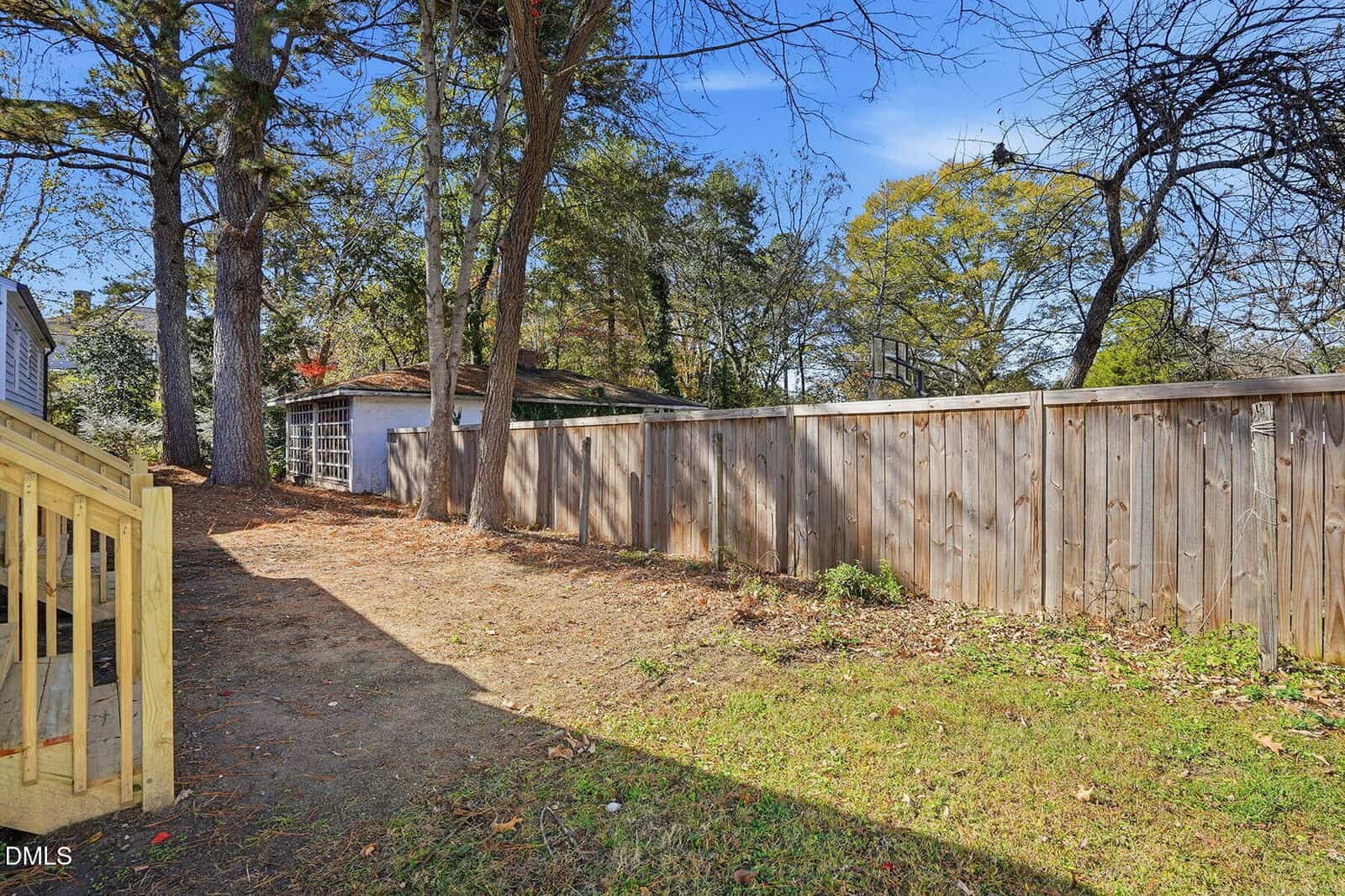 A backyard with sparse grass, bordered by a wooden fence and tall trees. There’s a small deck on the left and a detached garage in the background under a blue sky.