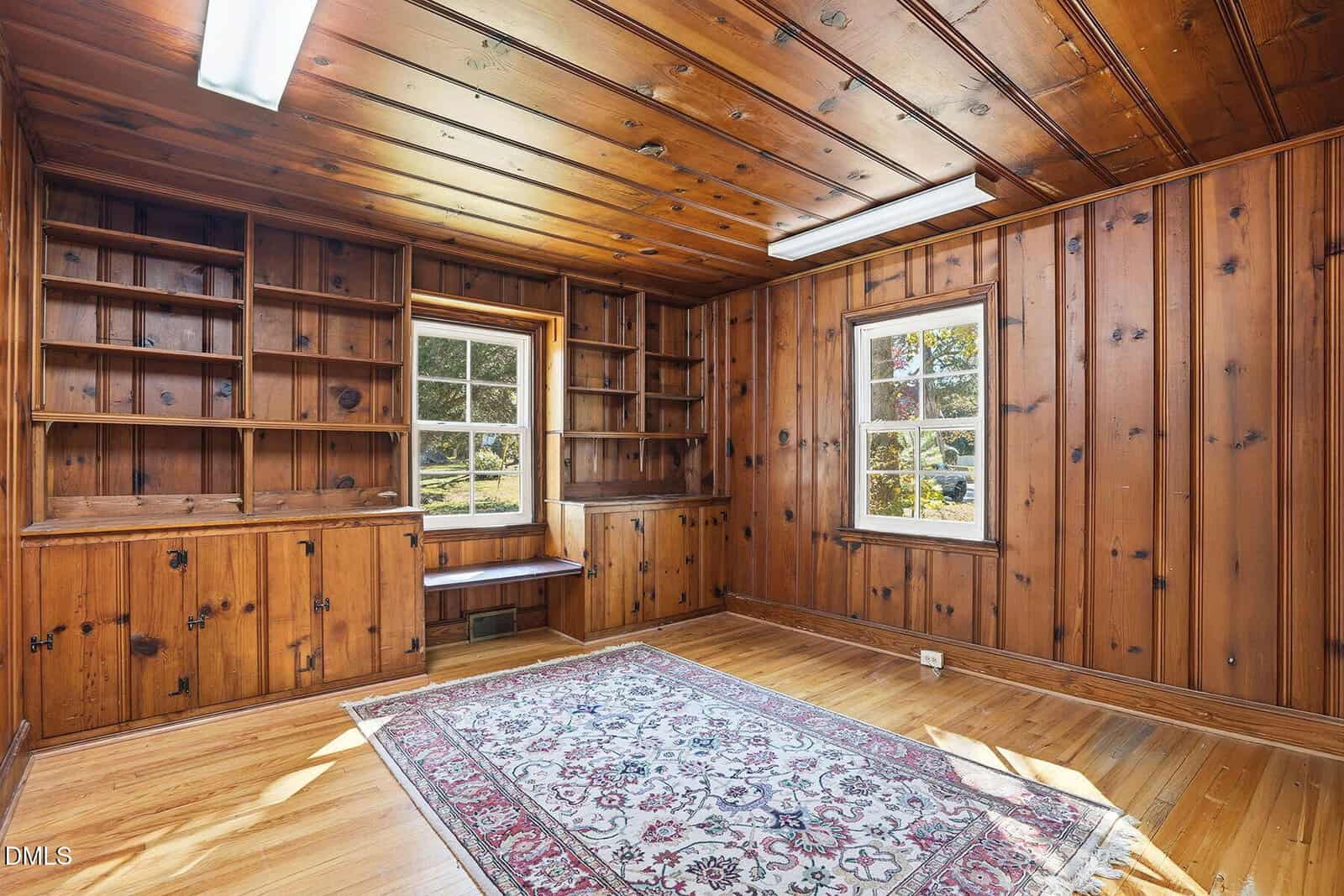 A wood-paneled room with built-in bookshelves, two windows, a window seat, fluorescent ceiling lights, and a patterned area rug on a hardwood floor. The room is empty of furniture.