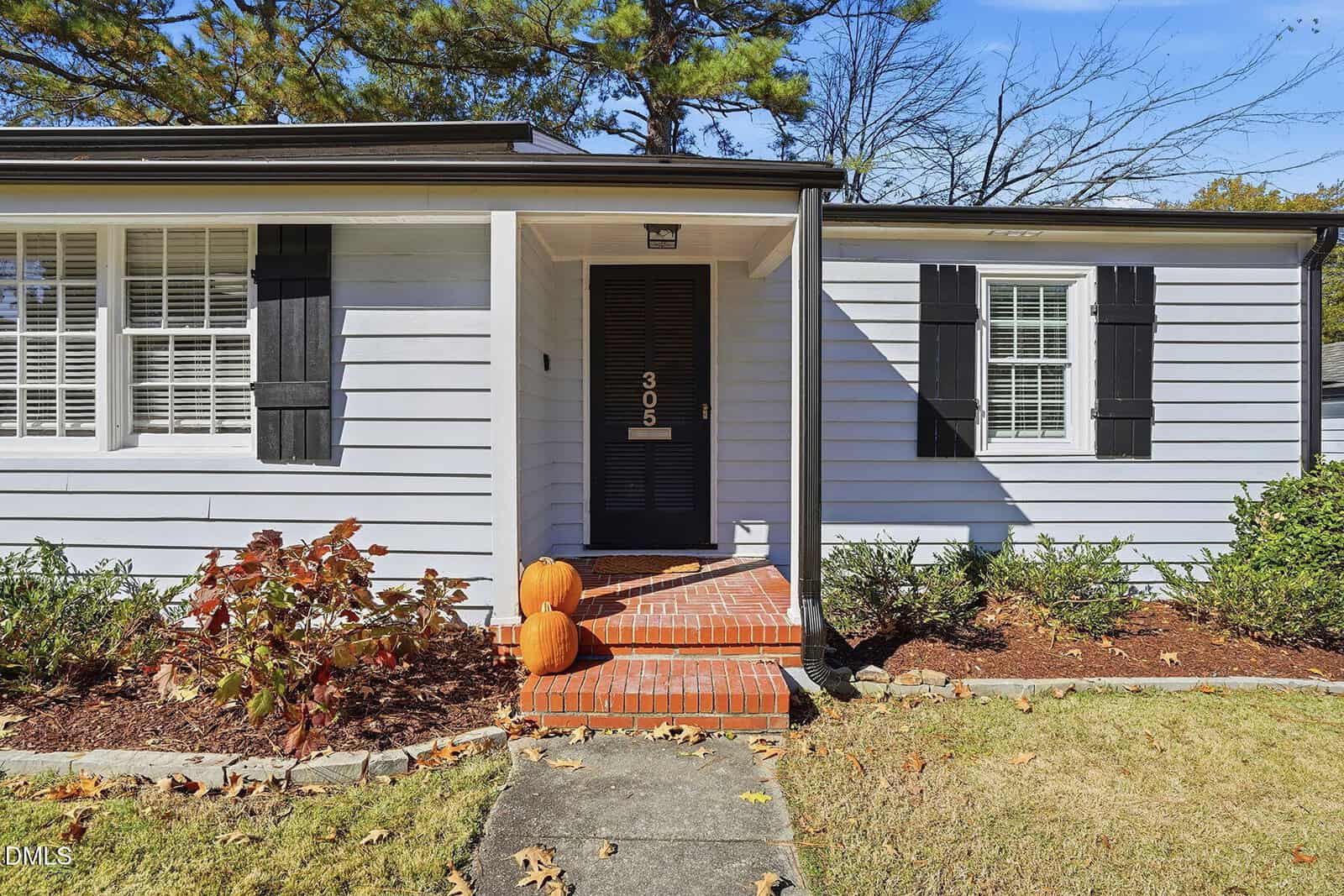 A small white house with black shutters and a black door marked 305. Red brick steps lead to the entrance, and two pumpkins are placed on the steps. Bushes and a lawn surround the walkway.