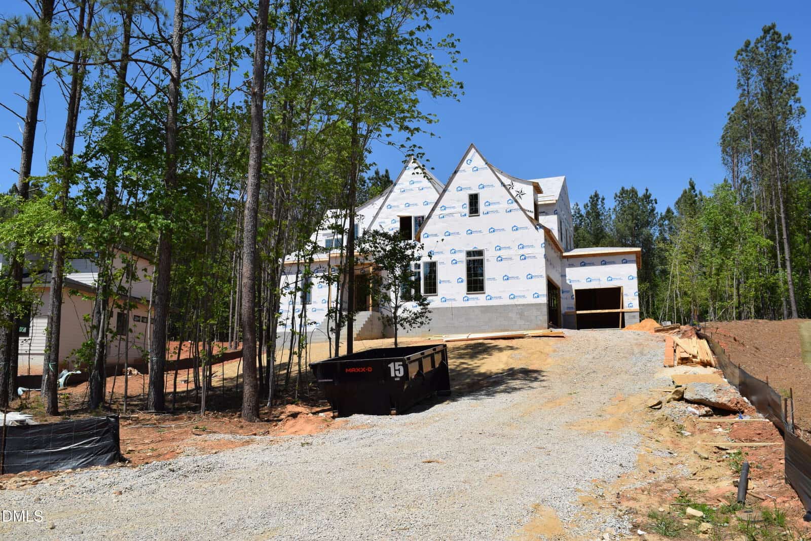 A two-story house under construction at 115 Spanish Oak Drive, Youngsville, features white sheathing and is surrounded by tall trees. A gravel driveway leads to the house, with a large black dumpster out front and blue sky over the woodland backdrop.