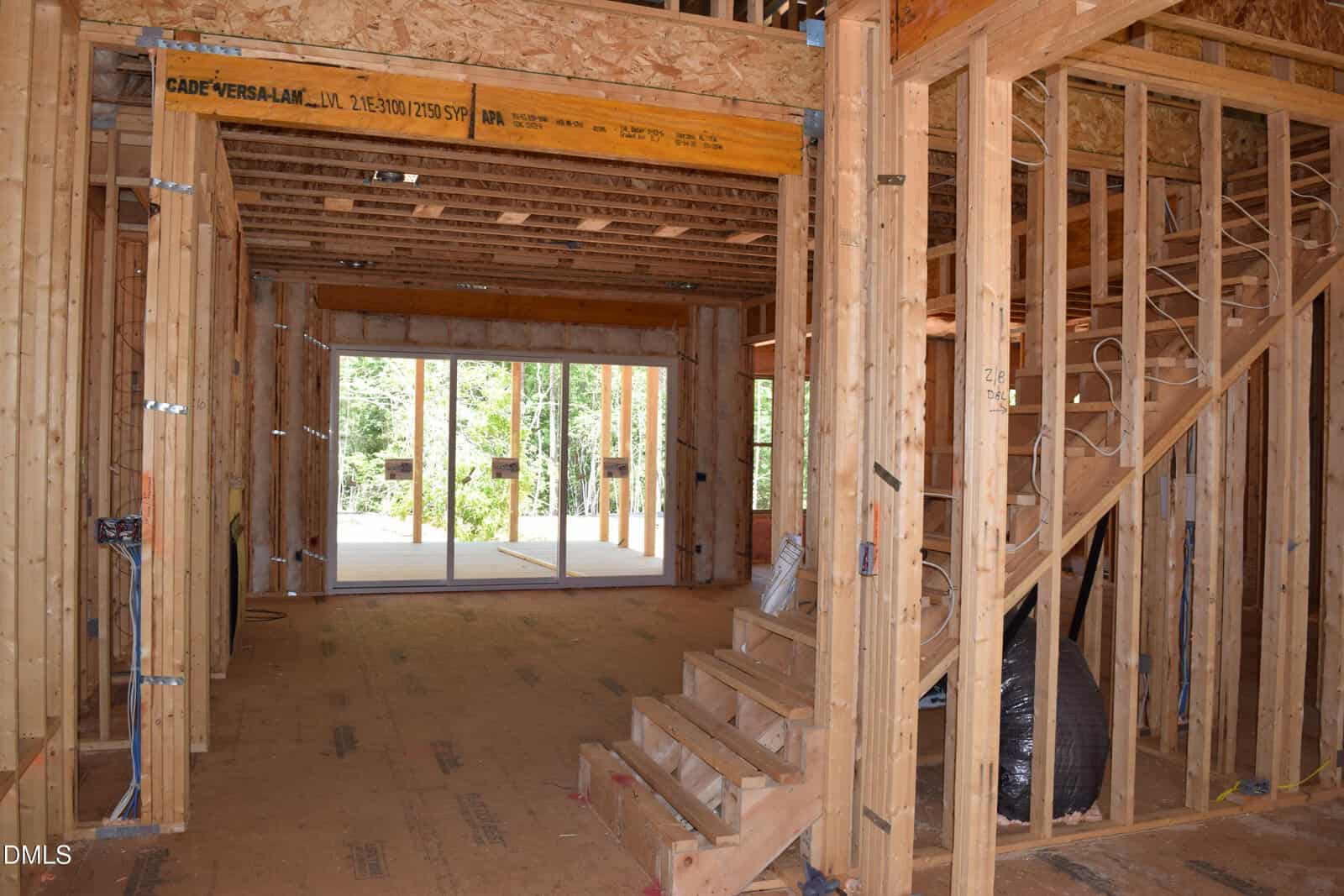 Interior view of a house under construction, showing exposed wooden framing, open stairs, visible wiring, and large sliding glass doors leading to a deck or outdoor area with trees visible outside.