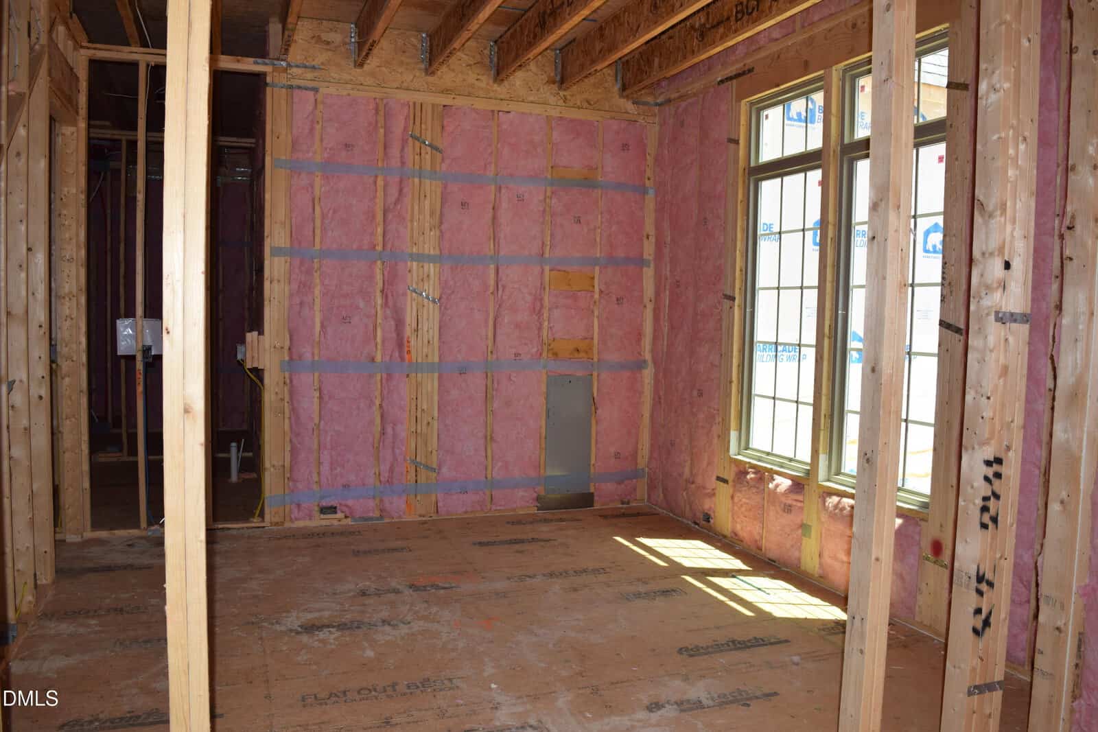 A room under construction with exposed wooden framing, pink insulation in the walls, and unfinished flooring. Sunlight enters through large windows on the right side of the image.