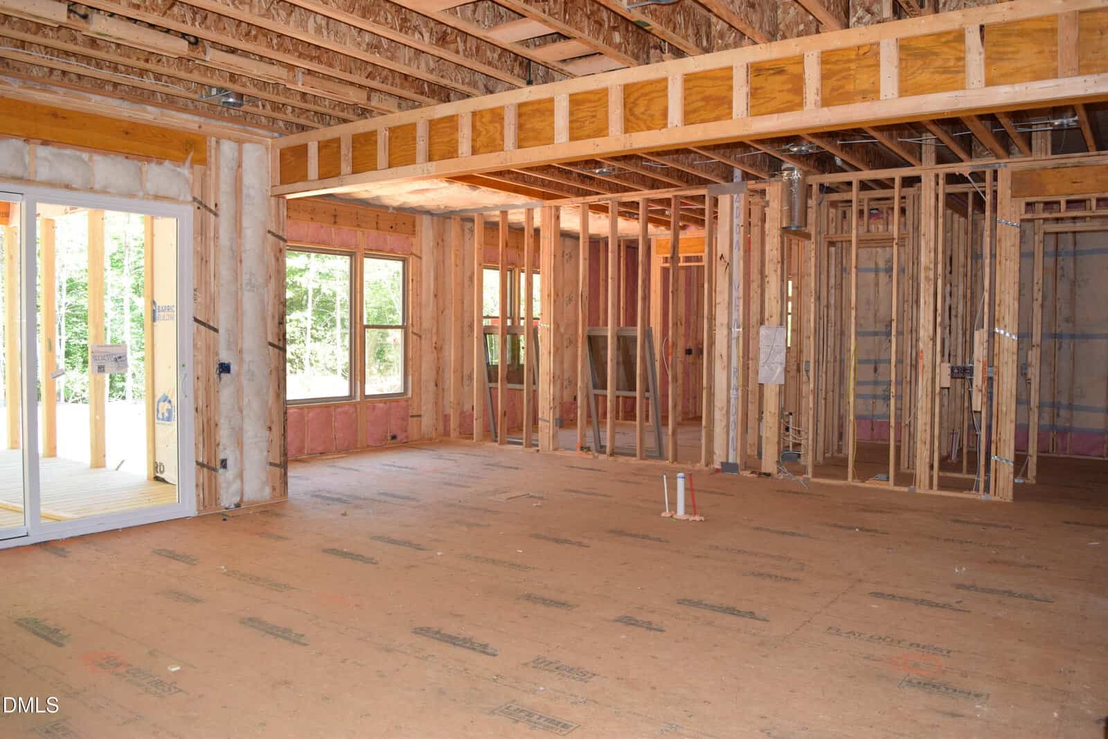 Interior of a house under construction, showing exposed wooden framing, ceiling beams, and unfinished walls. Large windows and a sliding glass door let in natural light. No drywall, flooring, or fixtures are installed yet.
