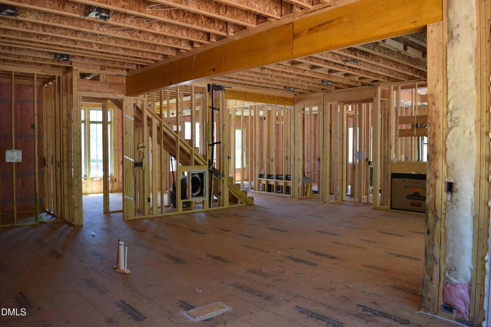 Interior view of a house under construction, showing exposed wooden framing, unfinished walls, visible insulation, and open ceiling beams. Stairs are partially built, and sunlight enters through window openings.